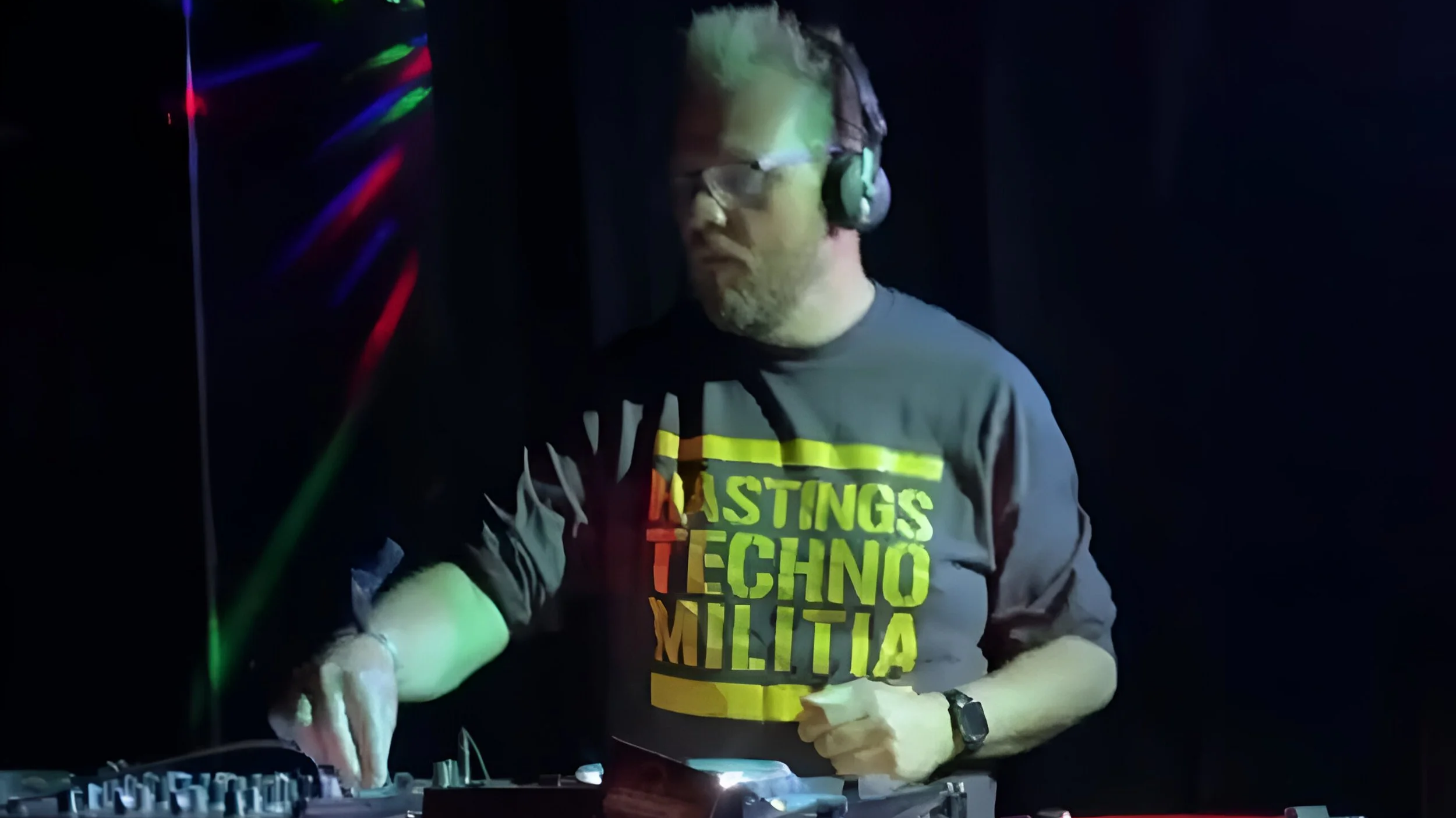 A man wearing glasses, headphones, and a black T-shirt with yellow, green, orange, and light green text that reads 'LASTINGS TECHNOLITA,' is DJing at a dark venue with colorful laser lights.