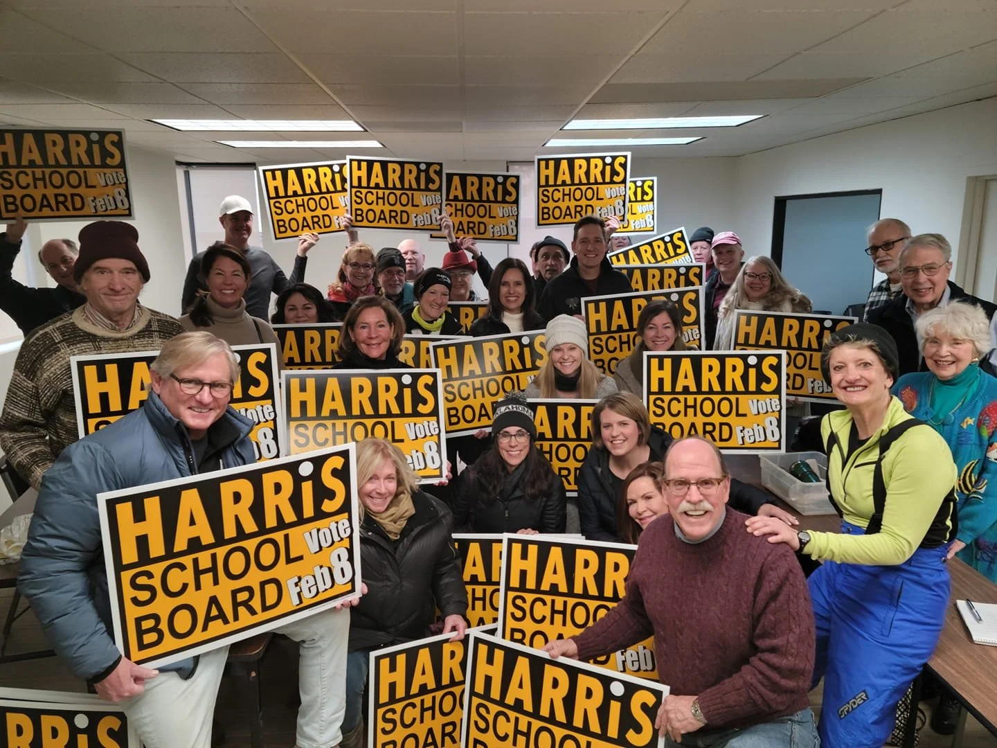 A group of people holding signs that say "Harris School Board Vote Feb 8." The image appears to be taken indoors, with participants smiling and posing together, suggesting support for a school board candidate.