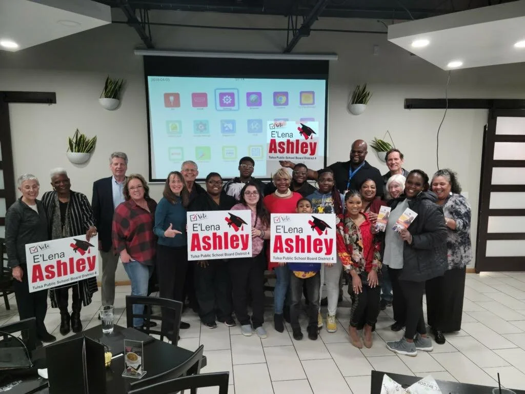 Group of people holding signs supporting E'Lena Ashley for Tulsa Public School Board District 4, standing in a room with a projector screen and plants on the wall.