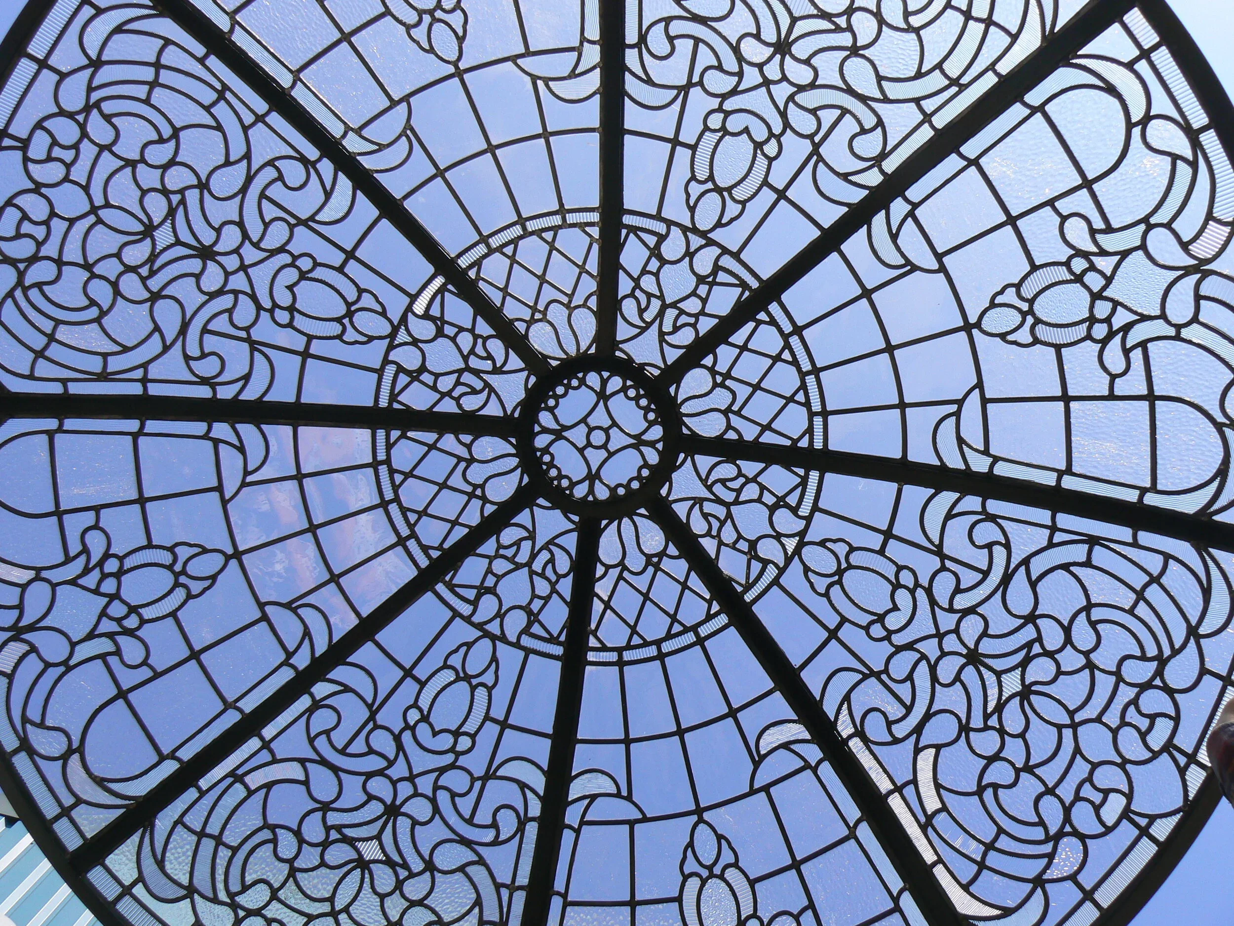 A stained glass dome ceiling with intricate swirling patterns and a view of the blue sky with some clouds through the glass.