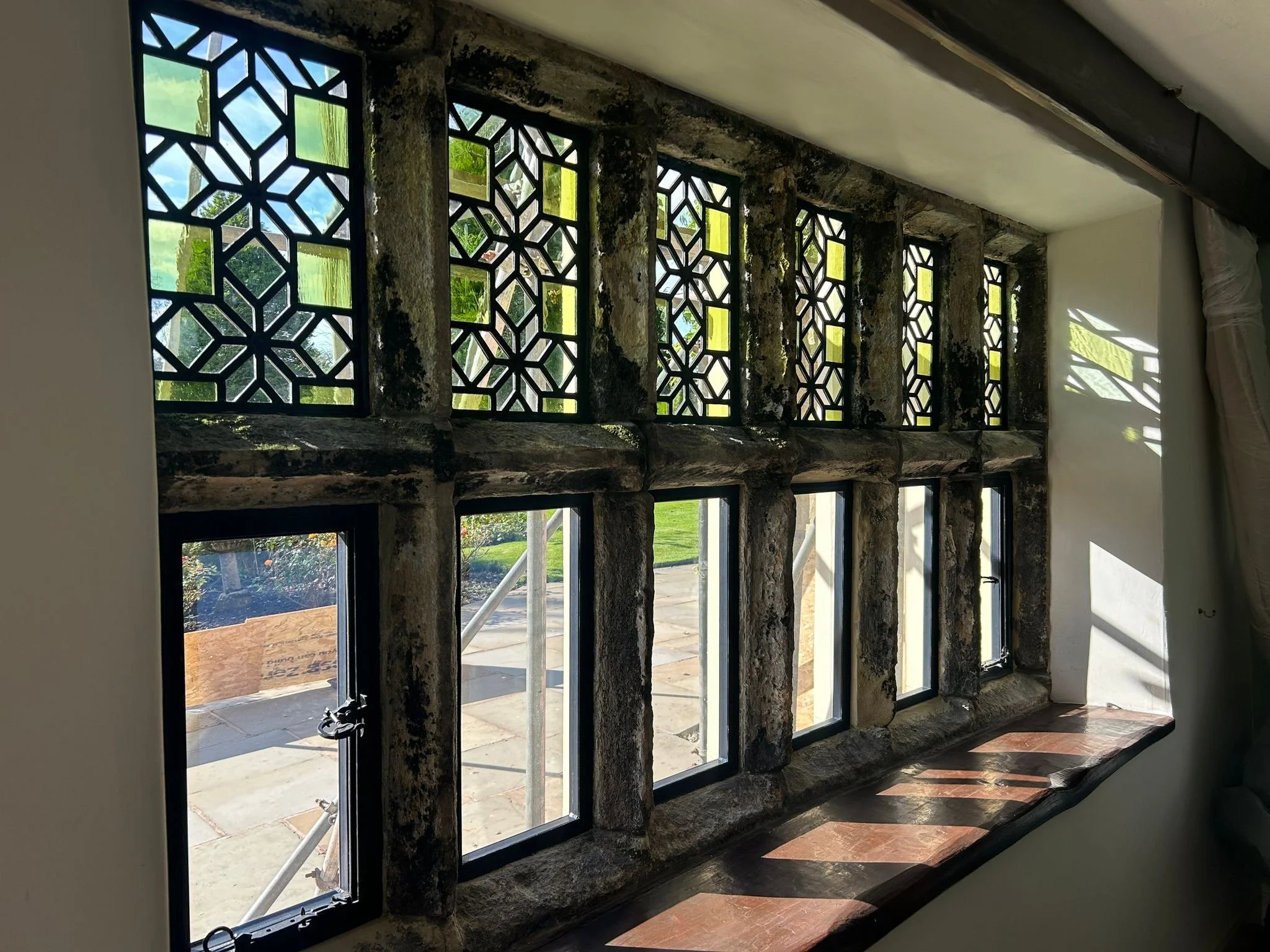 The image shows a window with decorative black metal grillework, located in a stone wall inside a building. Outside, there is a view of greenery and a paved patio area.