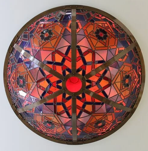 A decorative round wall clock featuring stained glass with geometric patterns in red, orange, purple, and black, and metal accents on a plain white wall.