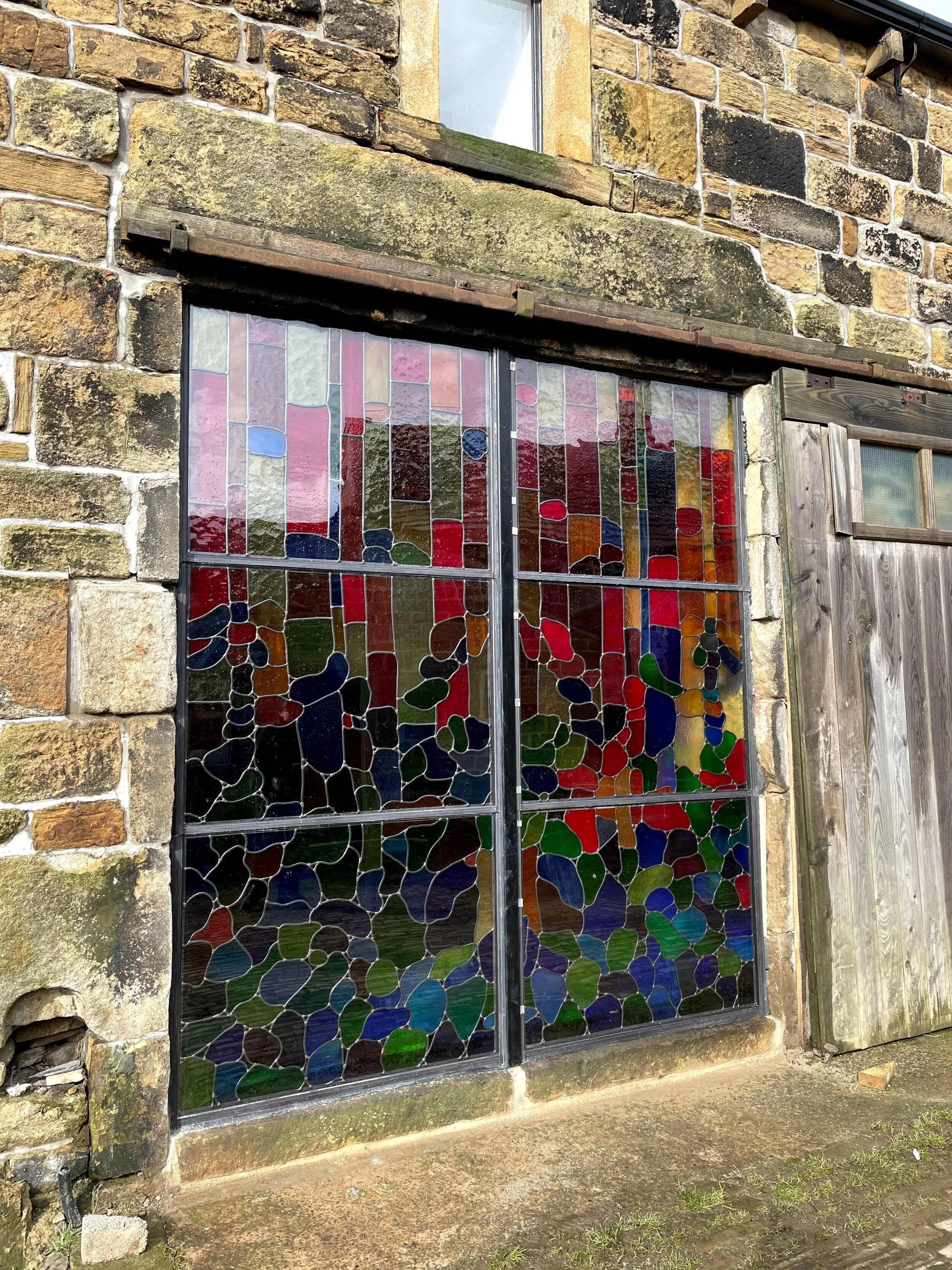 A window with a stained glass design of red, green, blue, yellow, purple, and orange pieces, set in a stone and wood wall exterior.