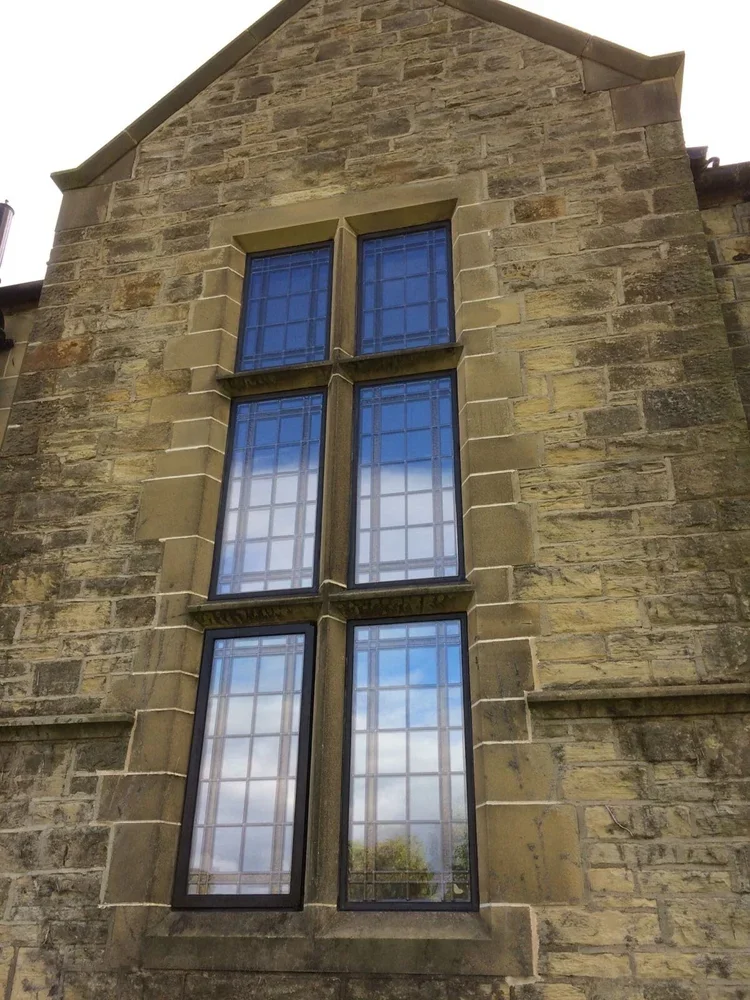 A stone building with a gothic-style arched window divided into six panes, with blue sky and clouds reflecting in the glass.