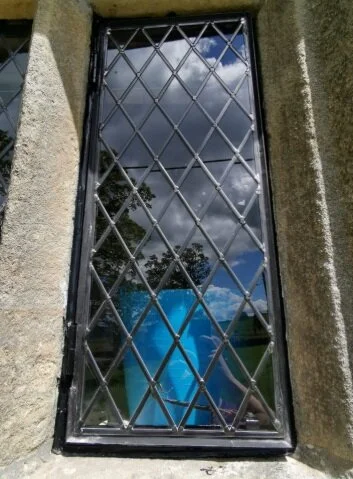 Reflective glass window with diamond-patterned security bars, surrounded by stone wall, reflecting cloudy sky and trees.