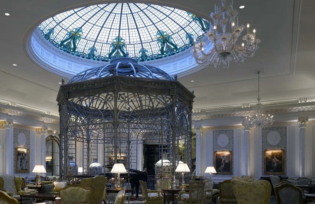 Elegant hotel lobby with ornate decor, chandeliers, a large glass dome skylight, and an intricate birdcage centerpiece.