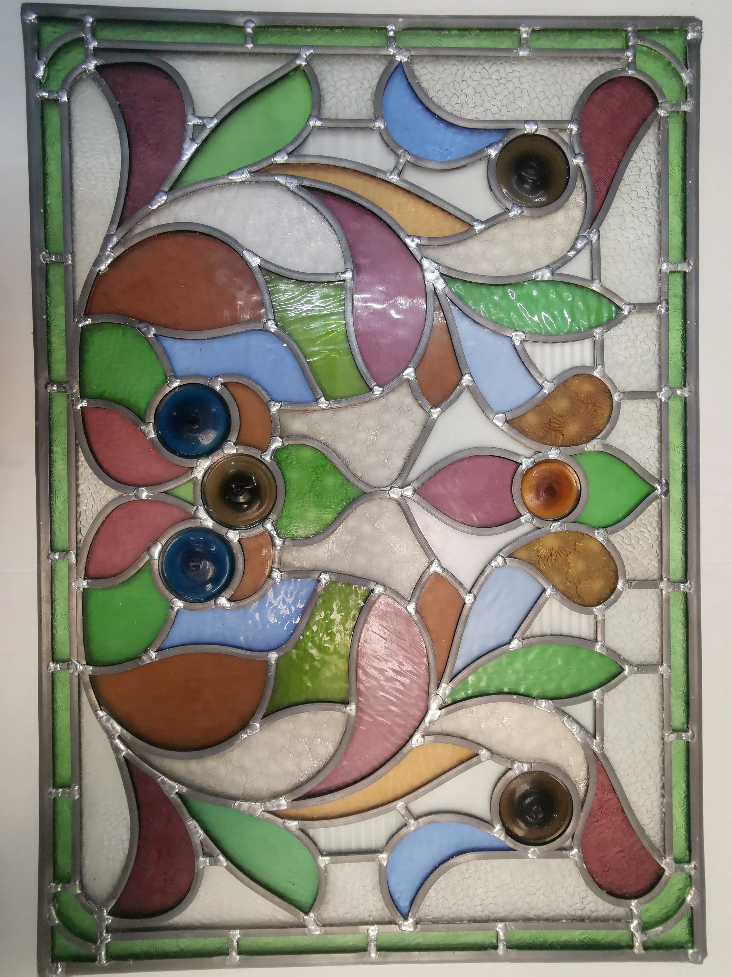 Colorful stained glass abstract design with various shapes and colors, including greens, blues, pinks, oranges, and browns, framed by a metal border.
