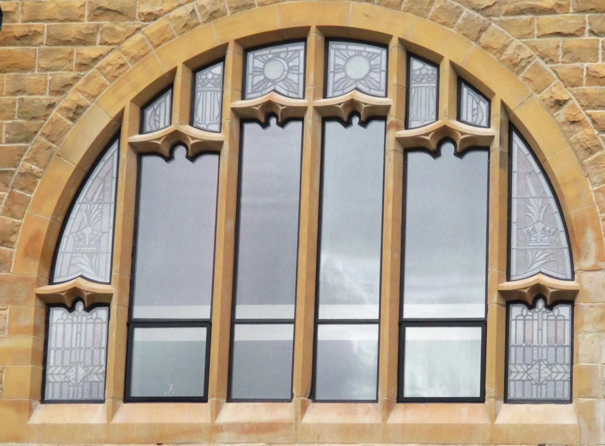 A large multi-pane stained glass window with decorative patterns, set in a brick wall with an arched top.