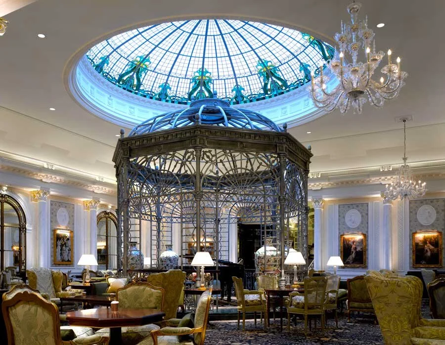 Elegant hotel lobby with a large glass skylight, ornate chandeliers, and a decorative iron structure in the center. The room has luxurious furniture and artwork on the walls.