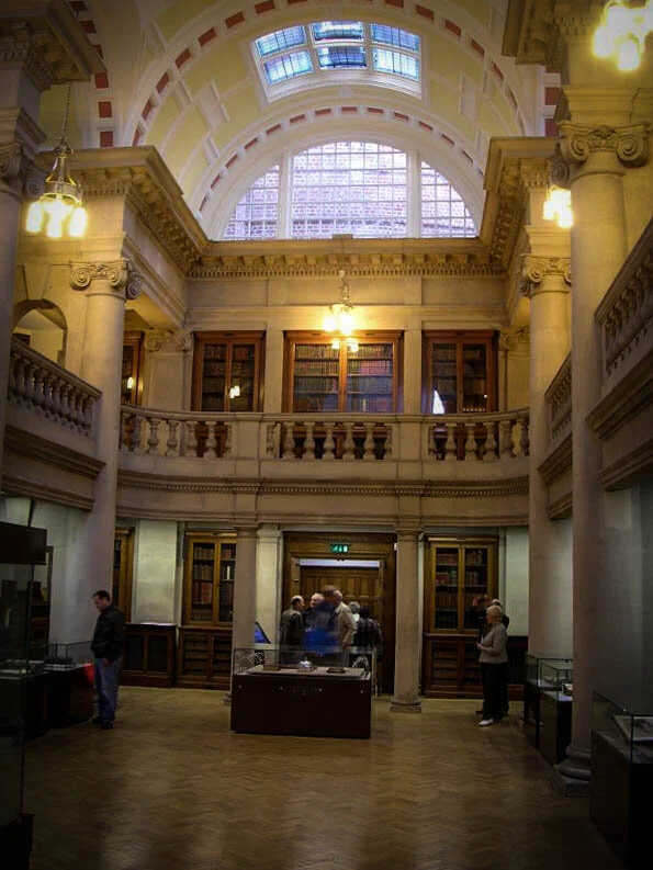 Interior of a historic library or museum with tall columns, wooden bookshelves, an arched glass ceiling, and several visitors viewing exhibits.