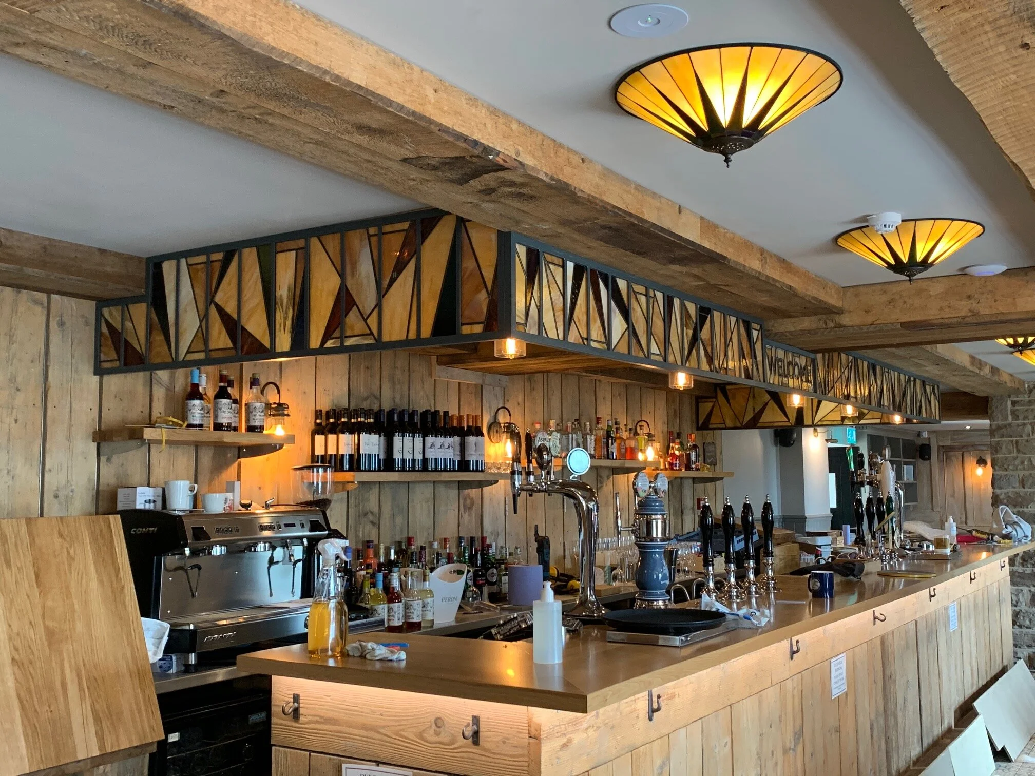 Wooden bar with beer taps, bottles of alcohol, and bar equipment under decorative stained glass fixtures on the ceiling.