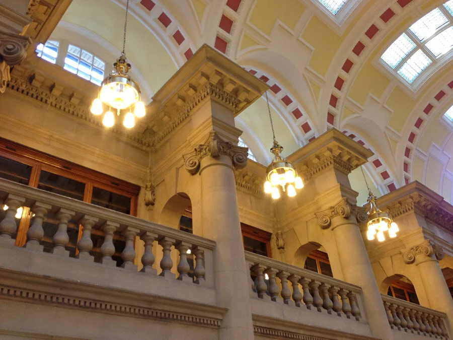 Interior view of a grand, classical building with tall columns, ornate ceiling with arches, and hanging chandeliers.