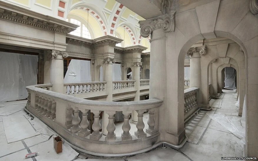 Interior of a historic building under renovation, featuring stone archways, columns, and decorative balustrades. Plastic coverings and construction materials are visible on the floor.