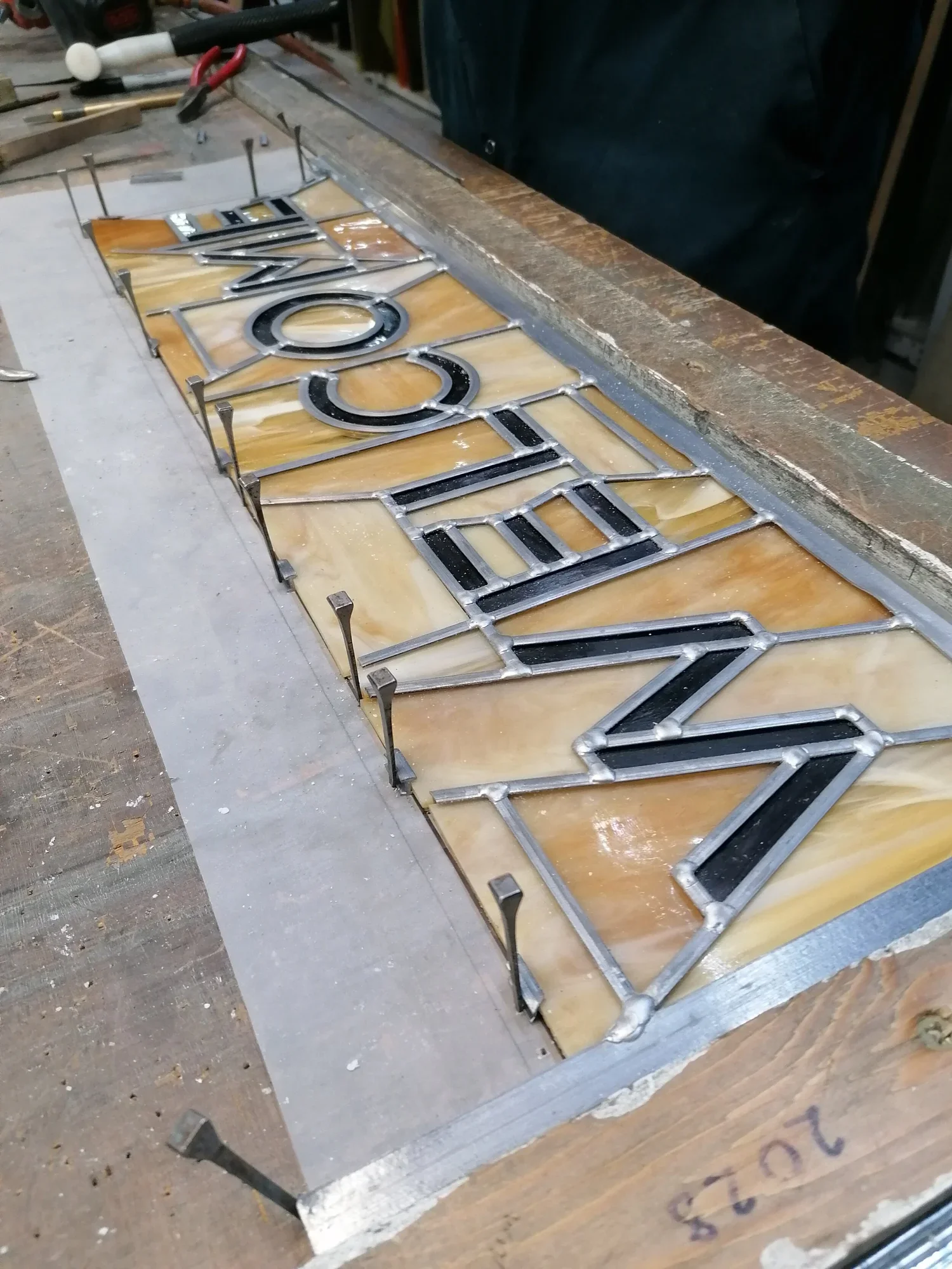 Close-up of a stained glass sign spelling 'WELCOME' with black and silver metal framing on a work table.
