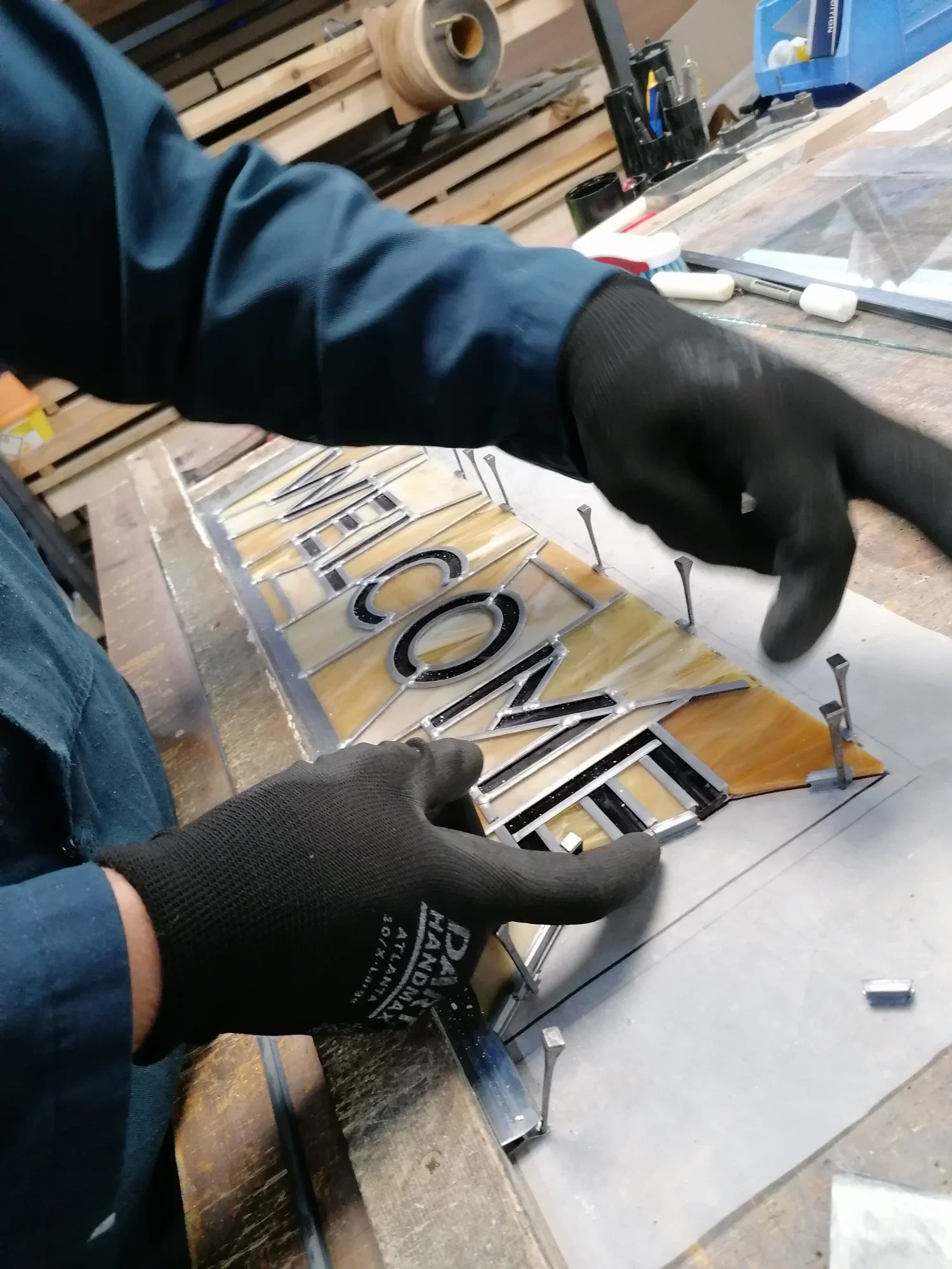 A person assembling a metal and acrylic sign that reads 'WELCOME' in a workshop, wearing black gloves.
