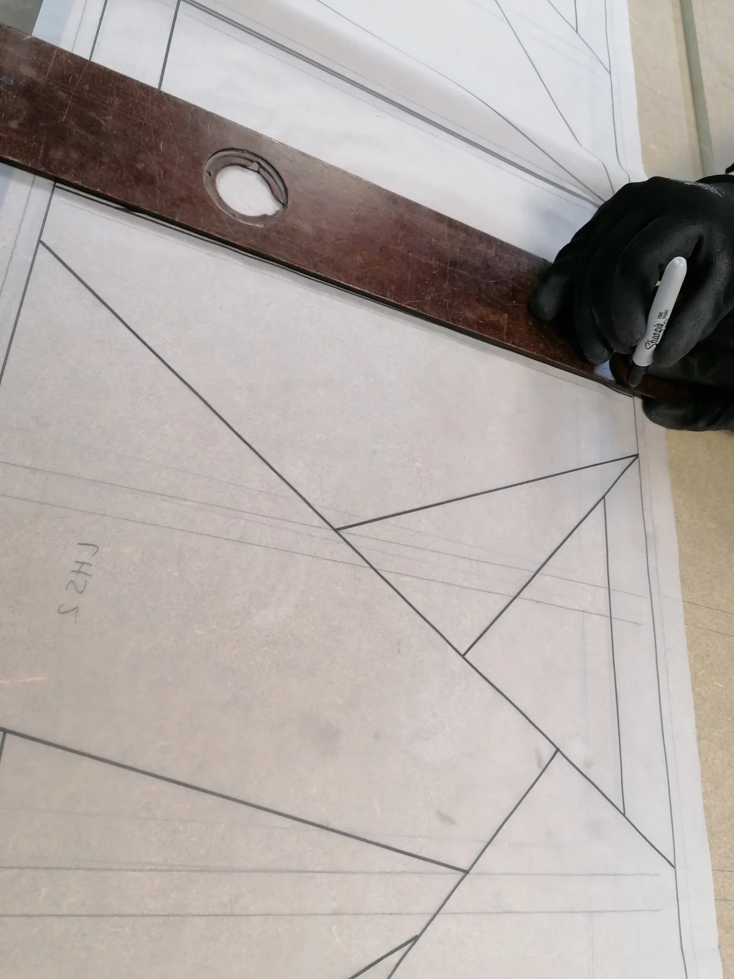 Person wearing black gloves is drawing geometric lines and triangles on a large sheet of paper or surface, with a ruler and a black Sharpie marker.