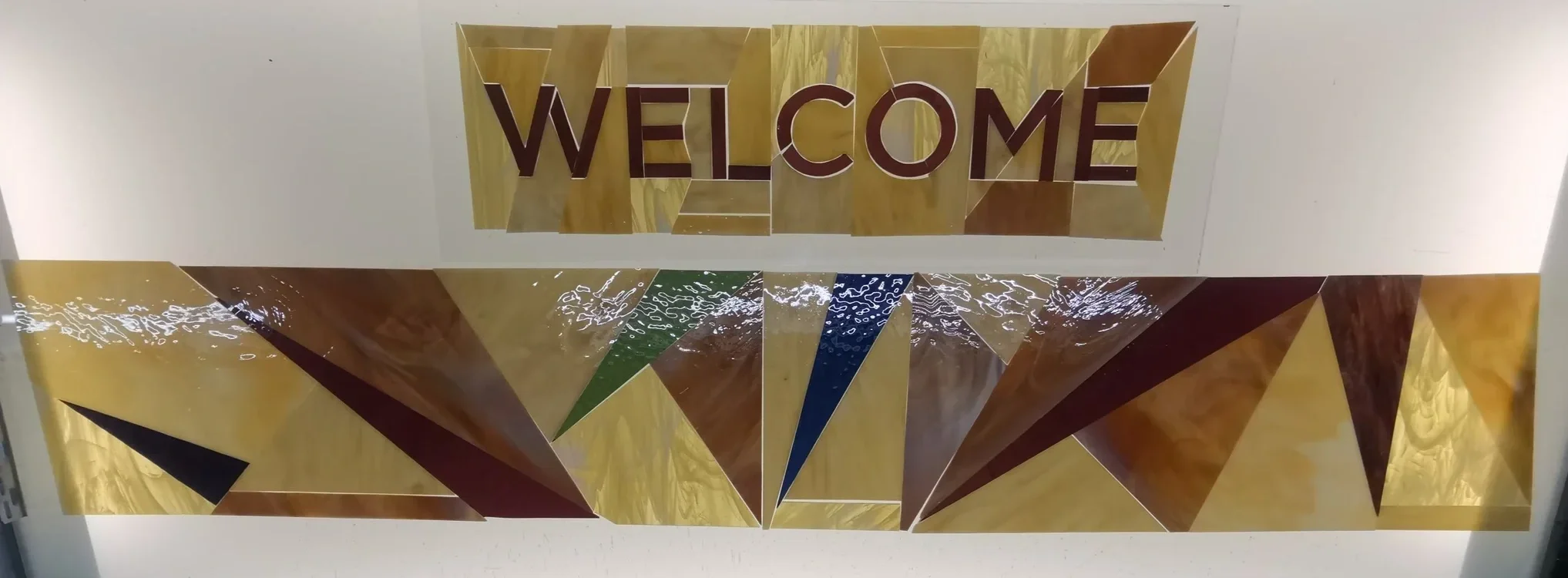 Decorative wall sign with the word 'WELCOME' in large letters, mounted above geometric patterns in yellow, brown, green, blue, and black on a white wall.