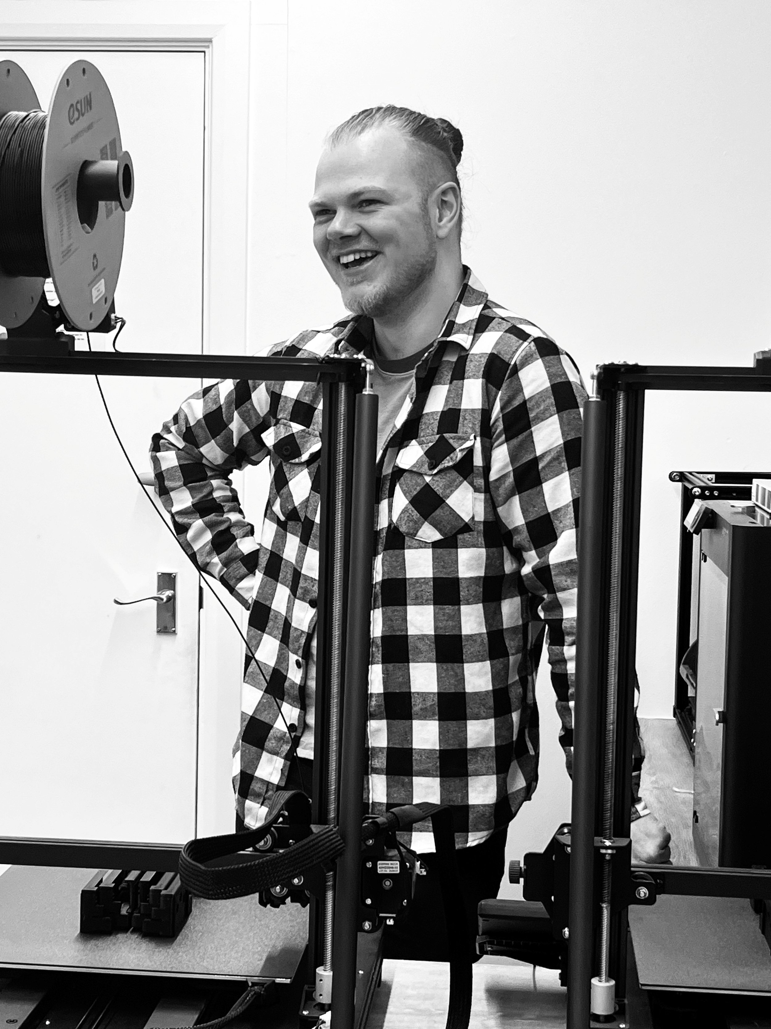 A man with a beard and long hair tied back, smiling while standing next to a 3D printer in a laboratory or workshop.