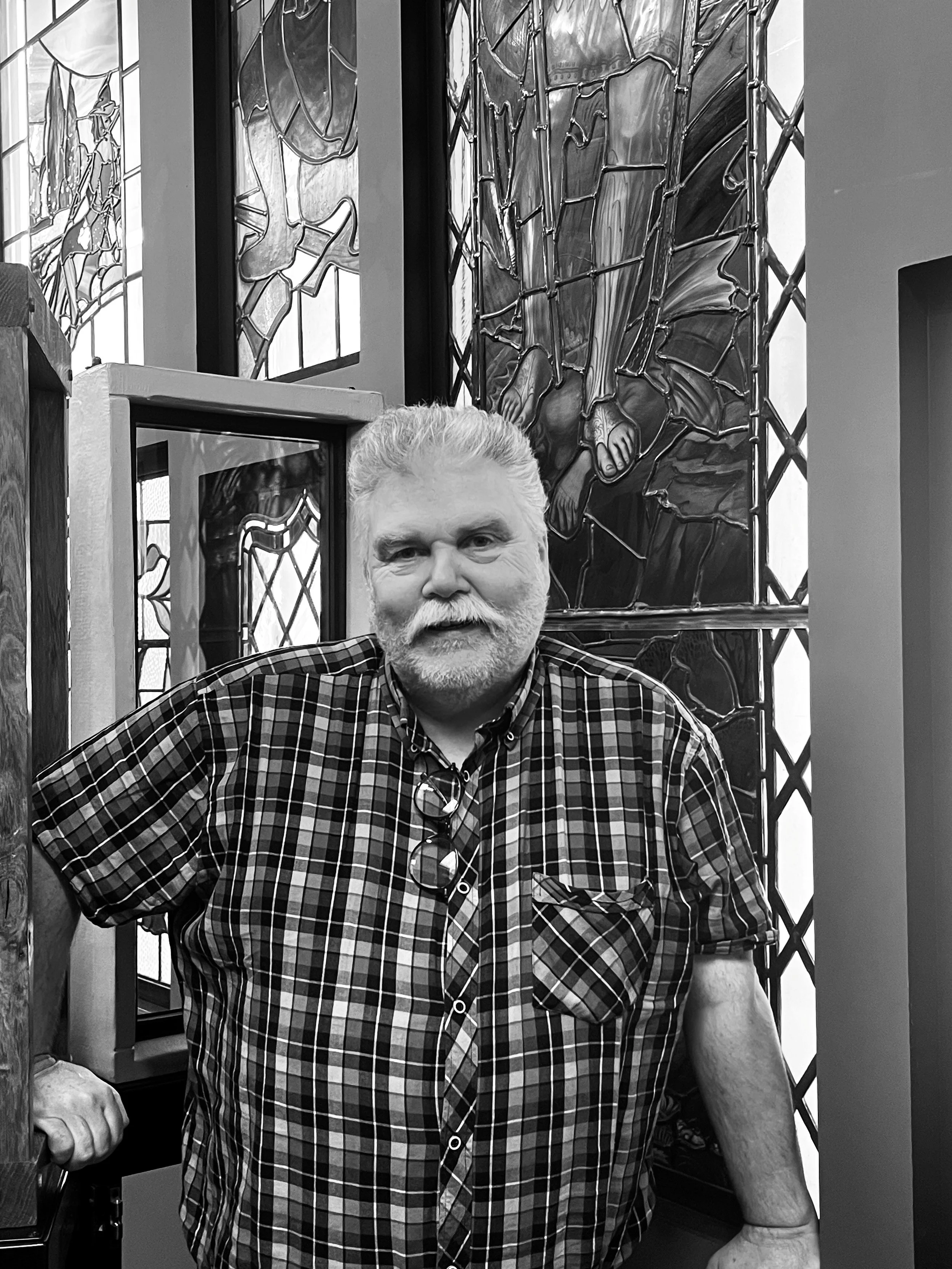 A man with a beard and glasses hanging from his shirt collar, wearing a plaid shirt, standing in front of stained glass windows depicting scenes with religious figures, inside a church.