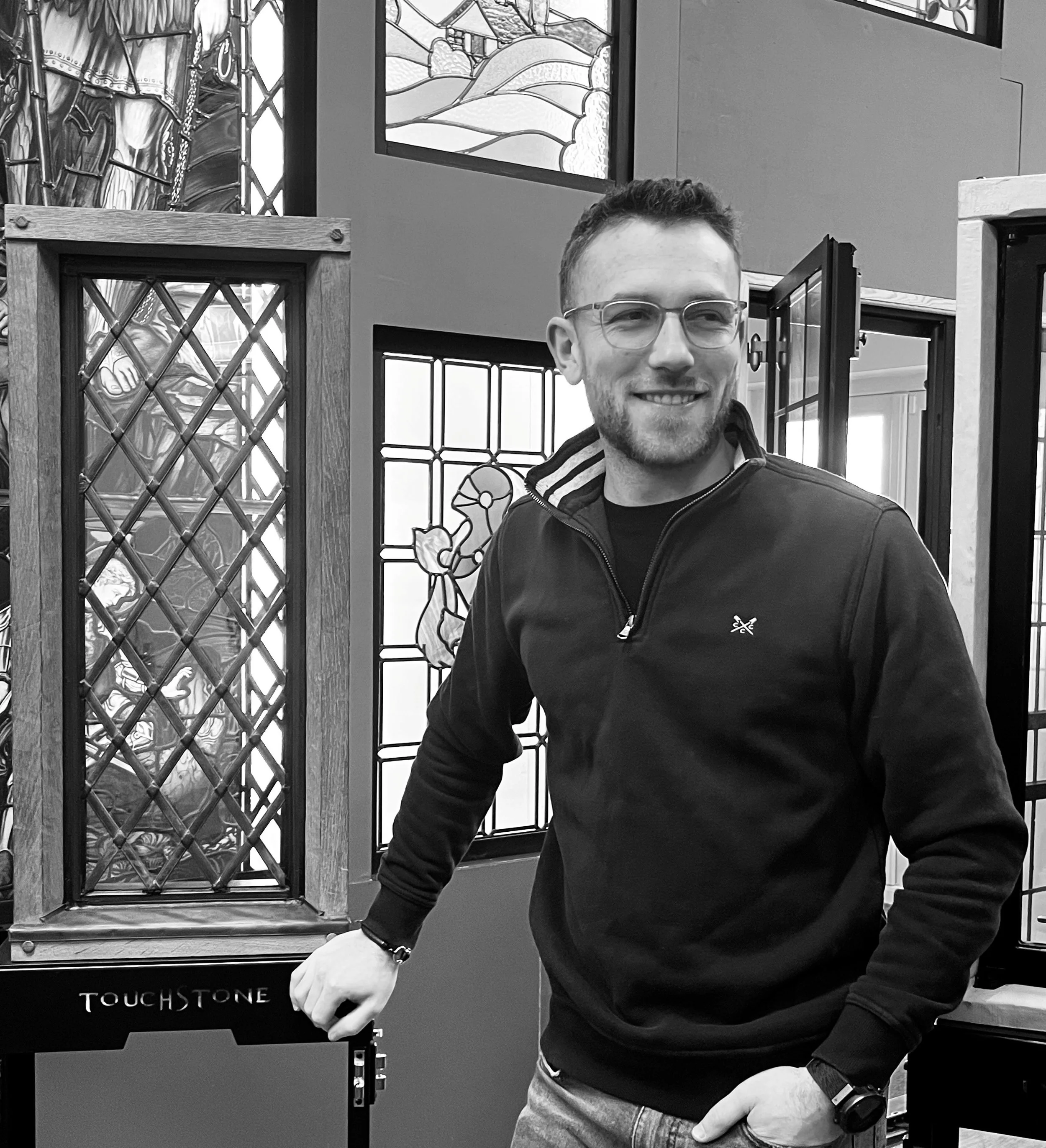 A smiling man with glasses and a beard stands indoors in front of stained glass windows and artwork, wearing a black zip-up jacket and jeans.