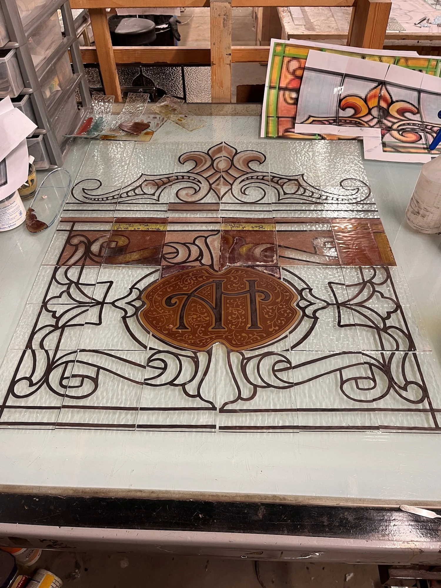 A glass worktable displaying a decorative tile design with intricate patterns and a central emblem featuring the letters "T" and "P," with various tile samples and tools around.