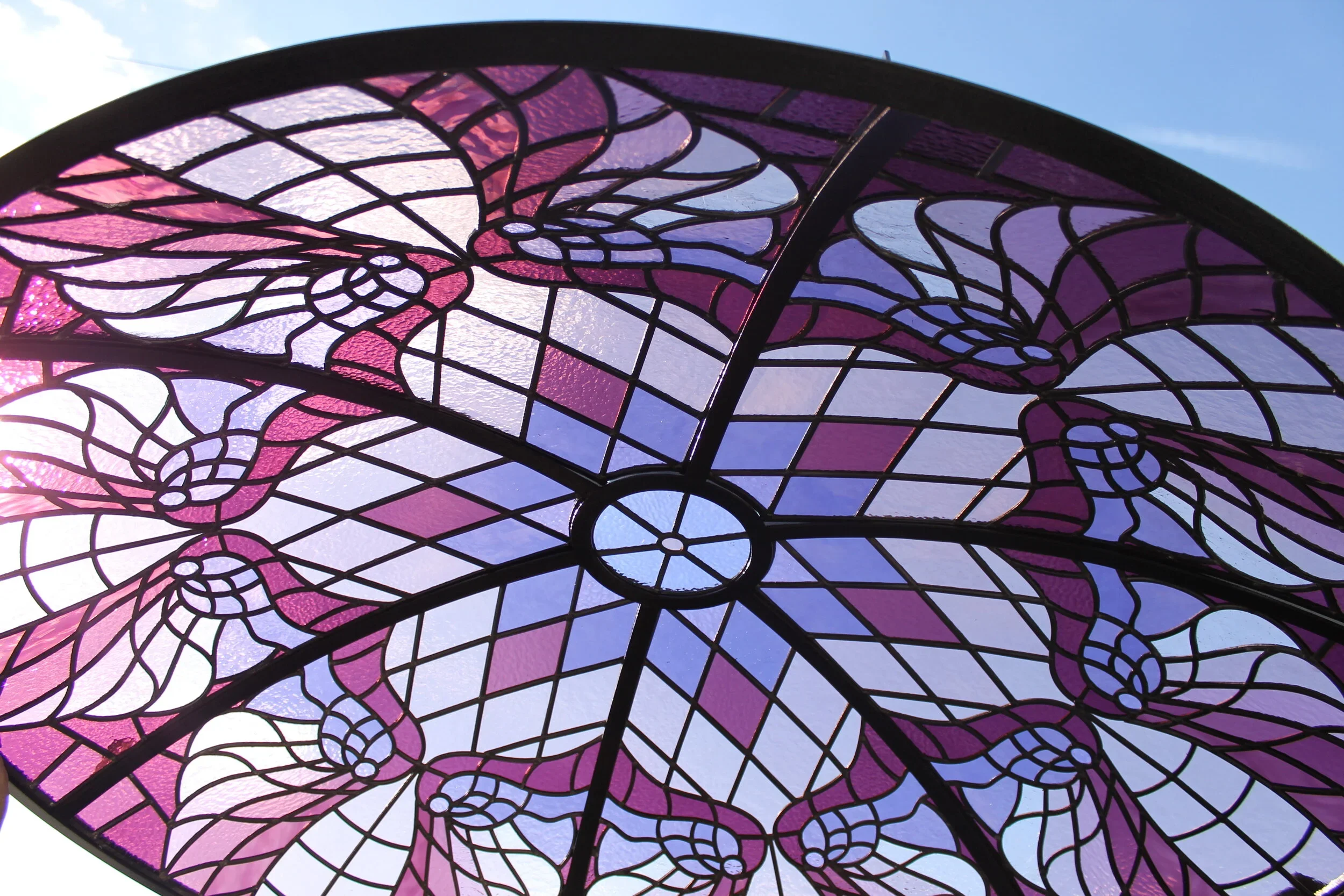 Close-up of a stained glass ceiling with purple and pink hues and intricate web-like design, set against a blue sky.