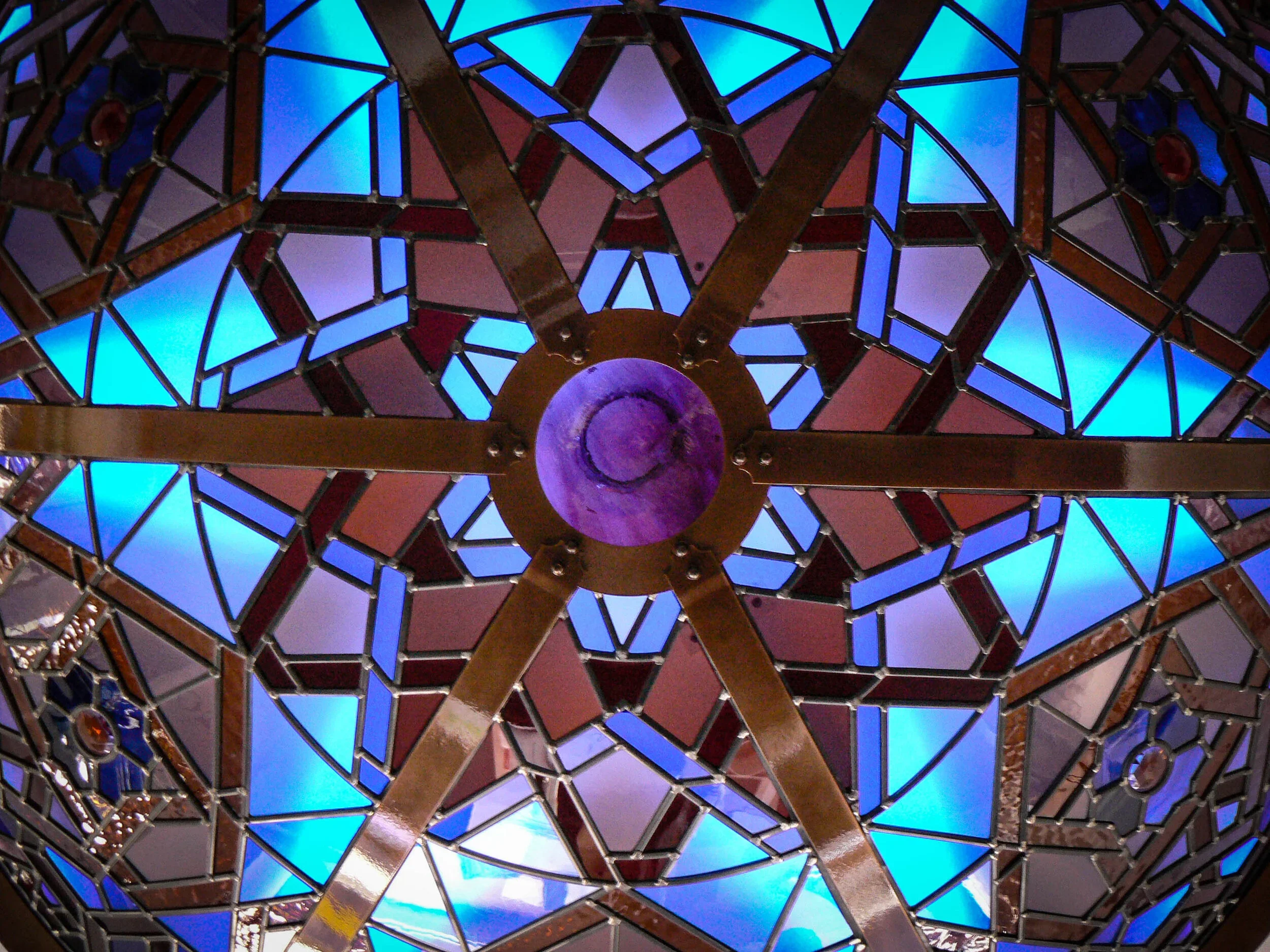 Close-up view of stained glass ceiling with colorful geometric patterns and metal framing, illuminated by blue light.