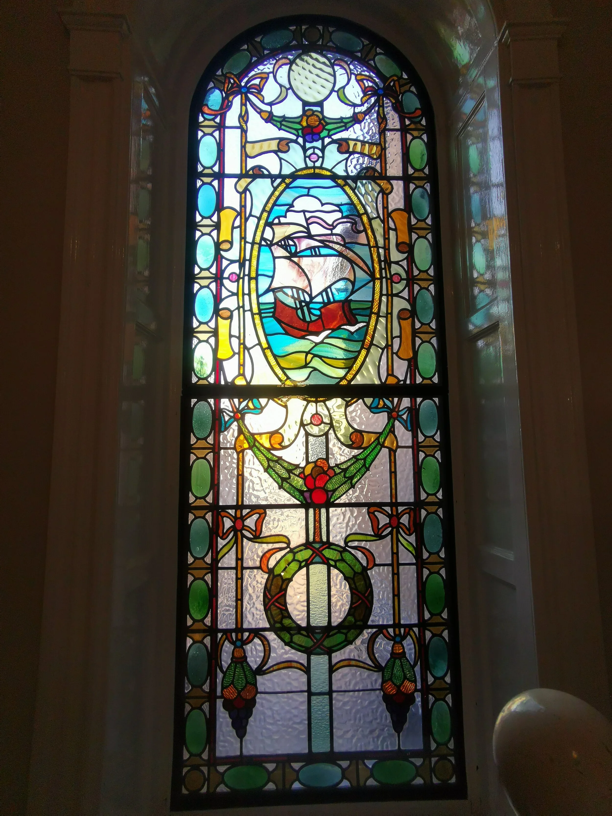 Colorful stained glass window featuring a central scene of a sailing ship on water, surrounded by decorative floral and geometric patterns.