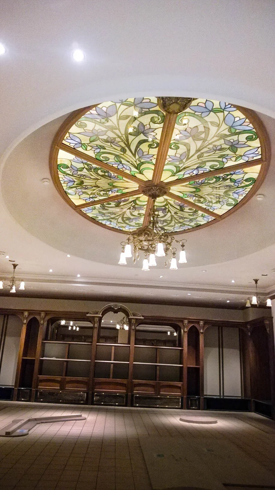 Interior room with a stained glass ceiling featuring floral patterns, ornate chandelier with multiple lights, and wooden shelving or cabinet.