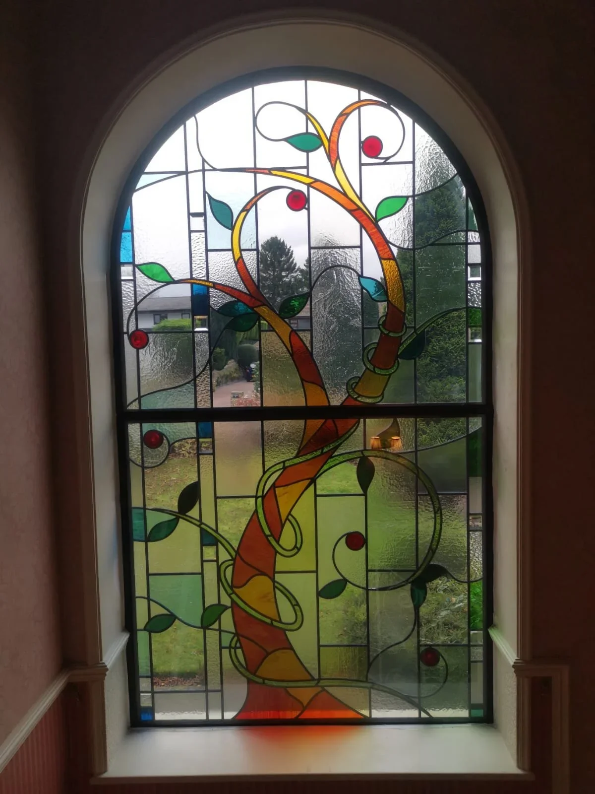 Stained glass window featuring colorful abstract design with swirling lines, leaves, and red circles, outdoors view visible through the glass.