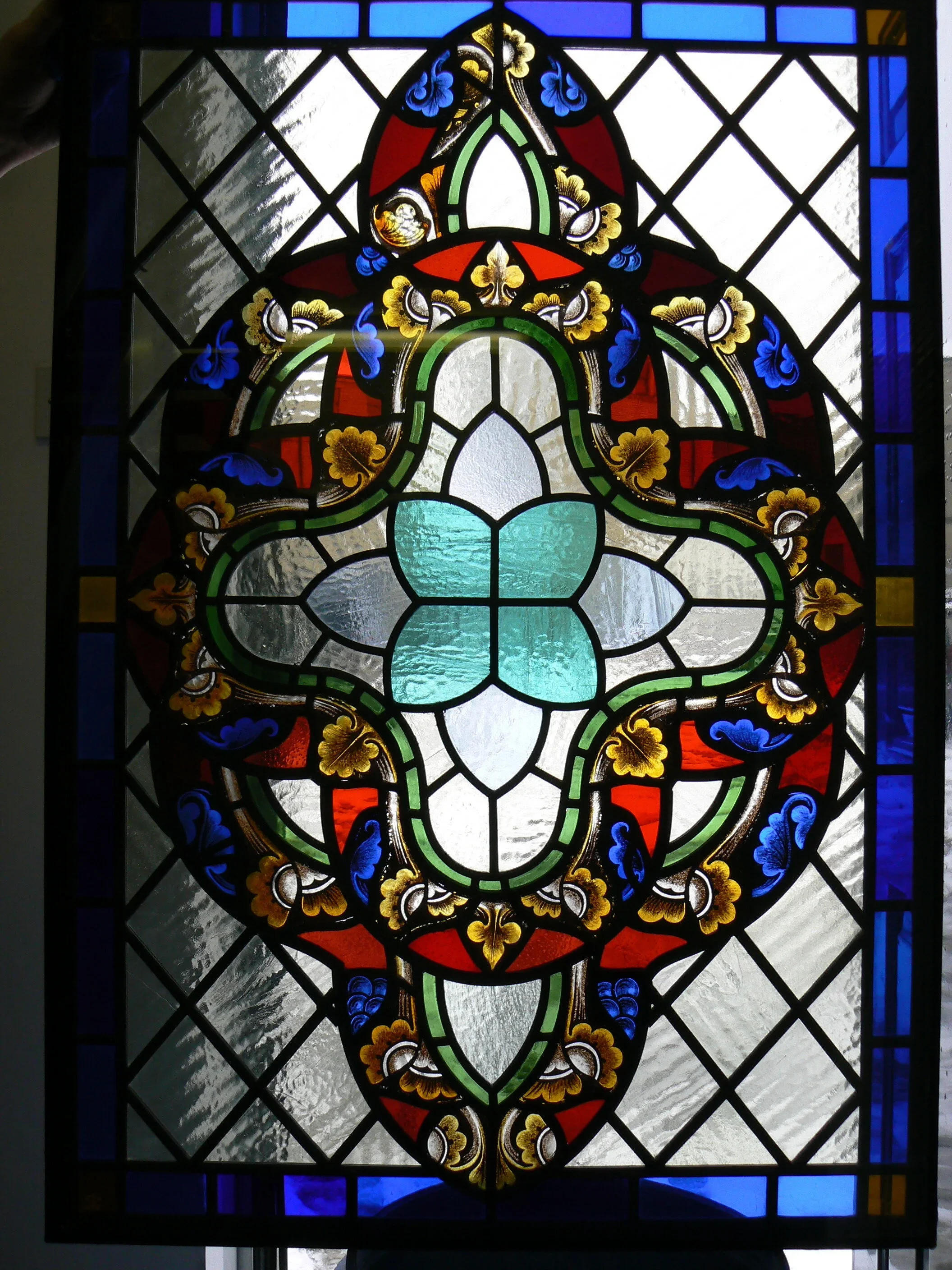A decorative stained glass window featuring an ornate pattern with red, blue, green, gold, and clear glass elements arranged in a symmetrical design.