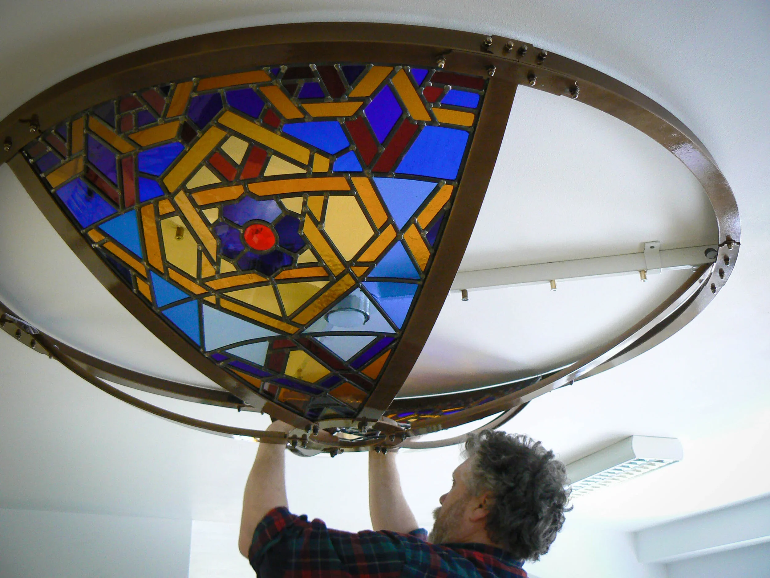 An elderly person with gray curly hair, wearing a plaid shirt, reaching up to adjust or work on a stained glass art piece on a ceiling, with white walls in the background.