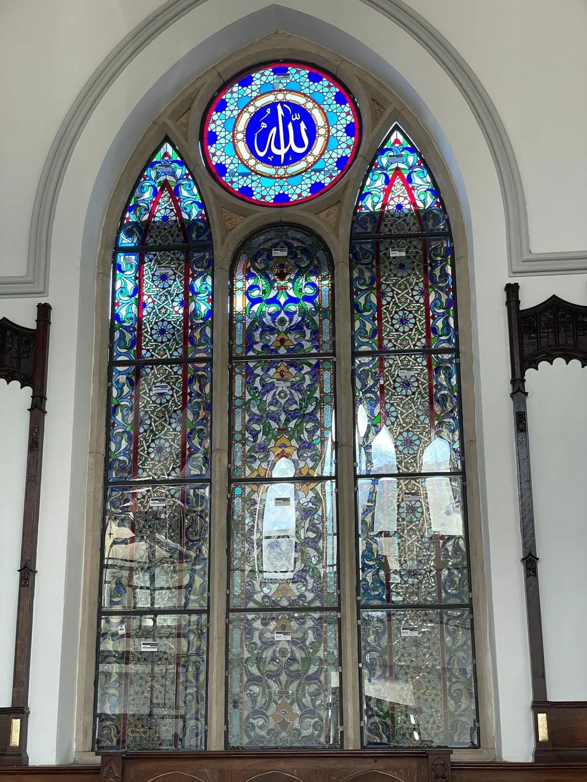 Colorful stained glass window in a mosque with intricate geometric and floral patterns. The top circular part features Arabic calligraphy with the word 'Allah' in white on a blue background.