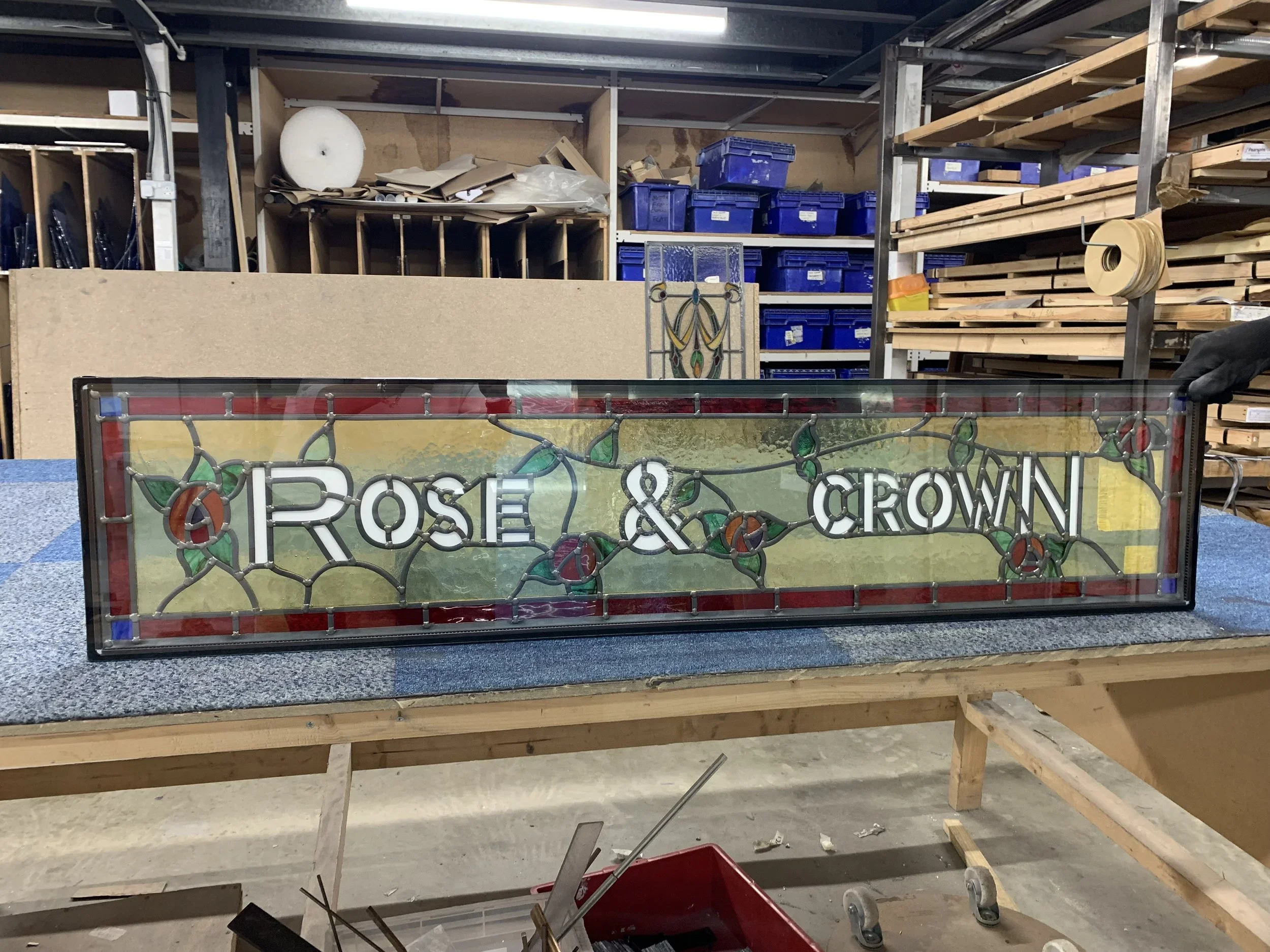 A woodworking workshop with shelves holding blue storage bins, wood planks, and a stained glass panel reading 'Positively Powered.'