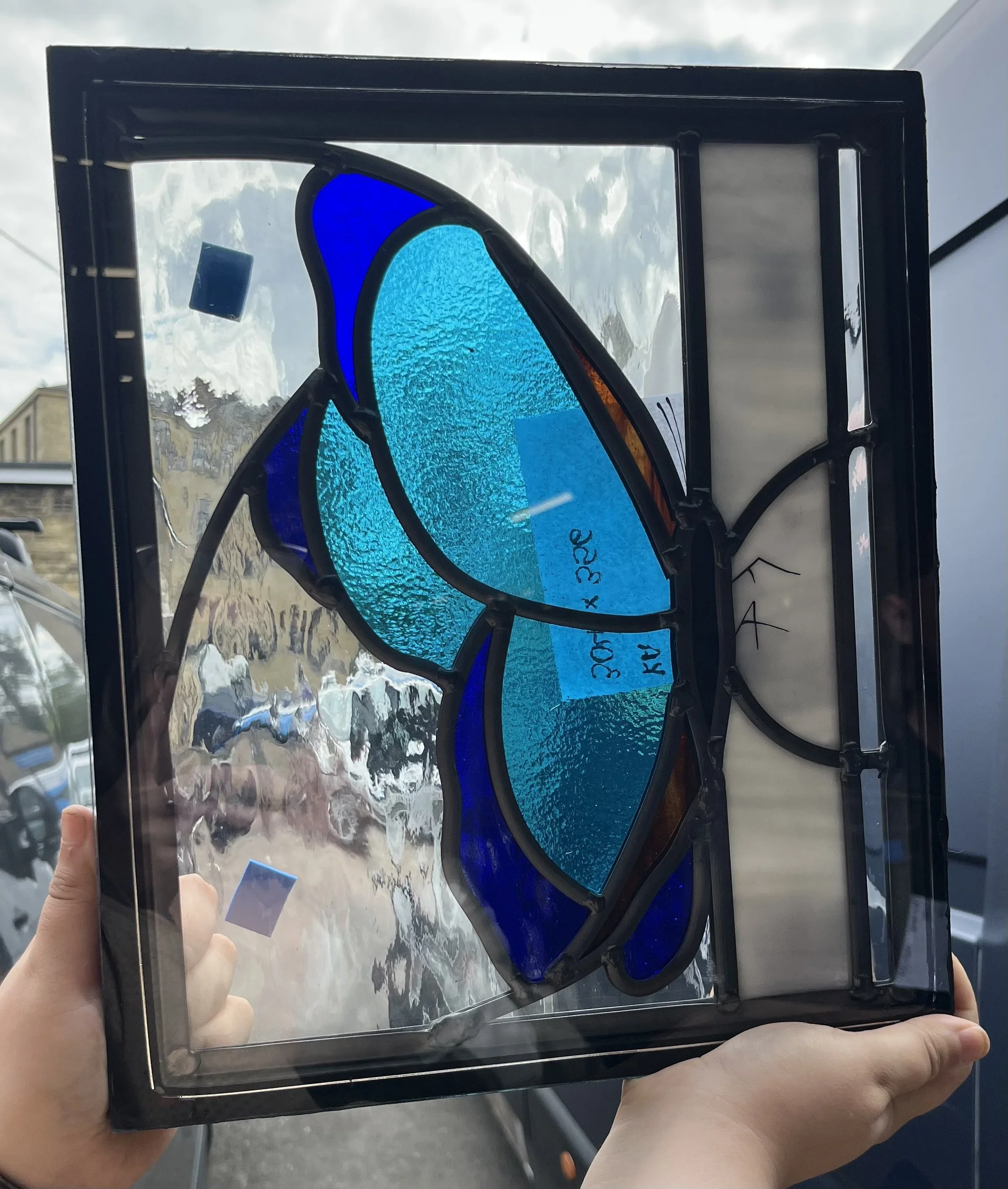 Colorful stained glass sunglasses with blue, teal, and amber sections held up against a cloudy sky and a person holding the frame indoors.