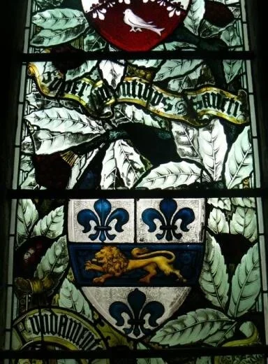 Stained glass window featuring a coat of arms with a shield, fleur-de-lis symbols, a lion, and Latin text, surrounded by a leafy design.