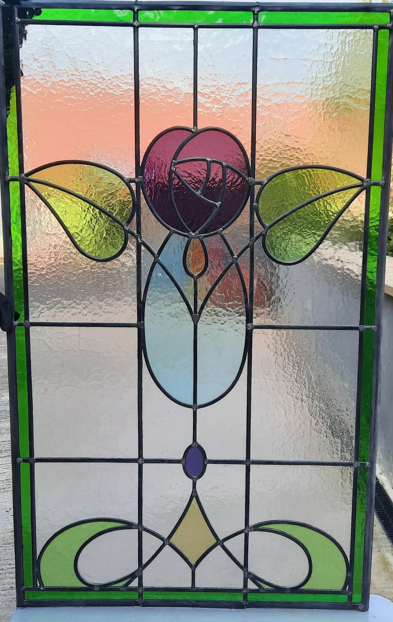 Decorative stained glass panel with a flower design featuring a purple flower at the top, green leaves on either side, and a yellow diamond shape at the bottom, framed with black metal and green borders.
