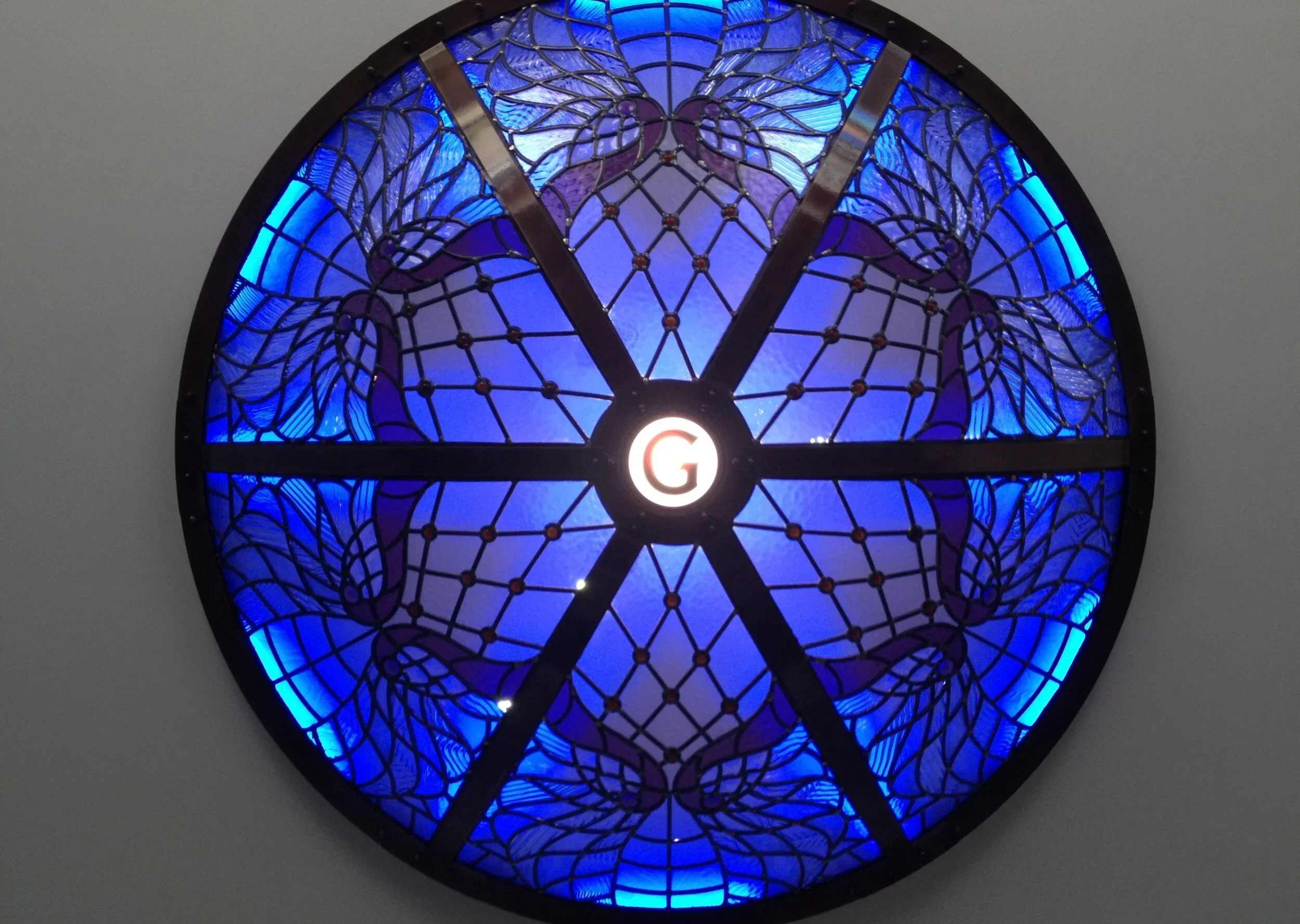 A round stained glass window with a butterfly design, predominantly in shades of blue and purple, with a white light at the bottom center.