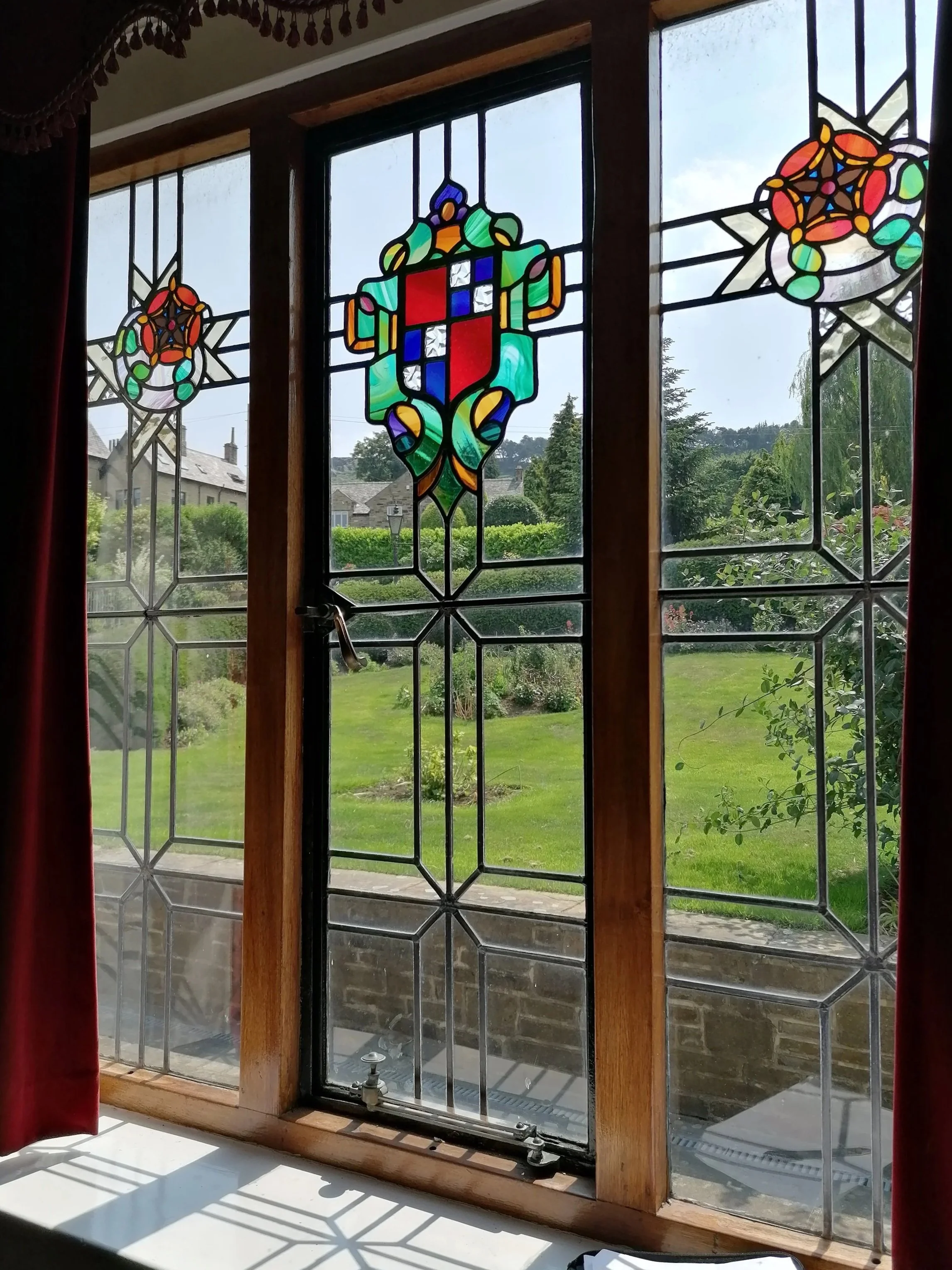 Stained glass window with a central shield and floral patterns, overlooking a green garden with hedges and trees.