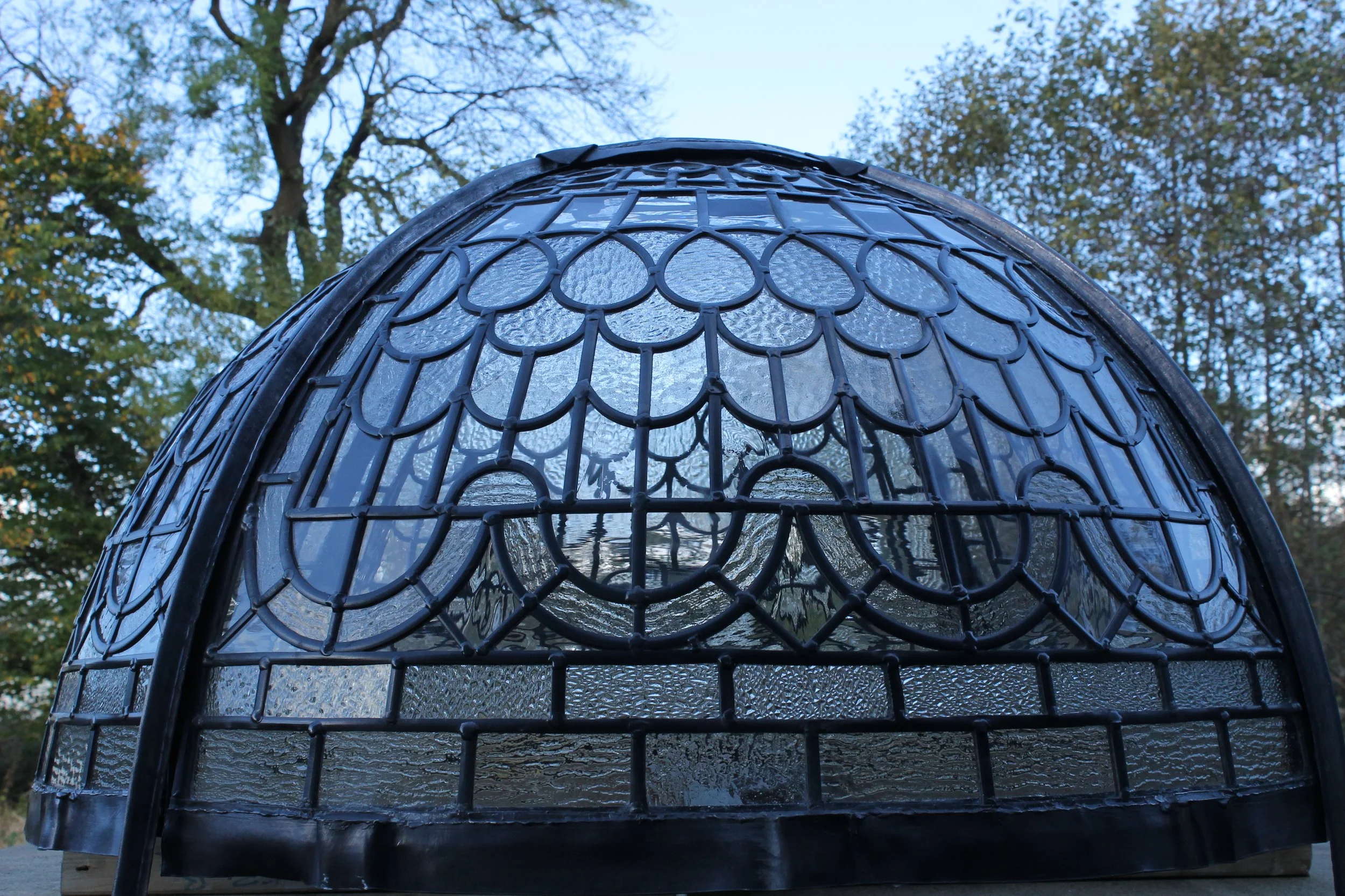 Close-up of a decorative stained glass lamp with intricate black framework and textured glass panels, outdoors with trees in the background.