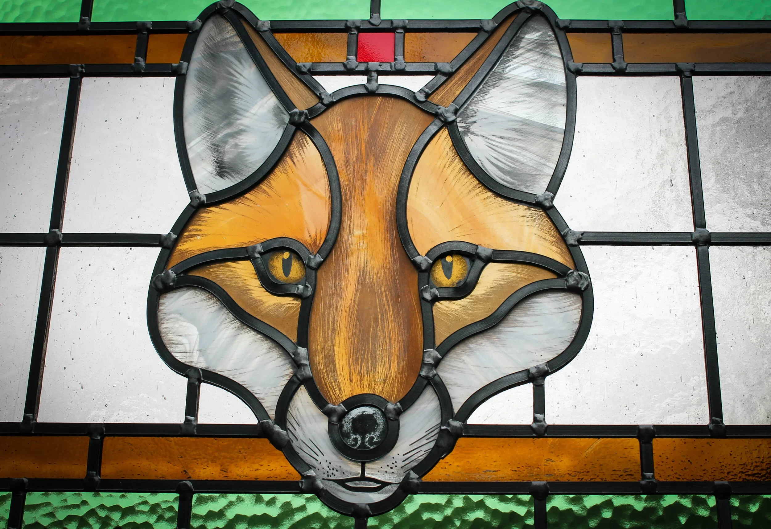 A stained glass panel featuring a fox's face, with orange, white, and black glass pieces, framed with black lead, and a red rectangular piece at the top.