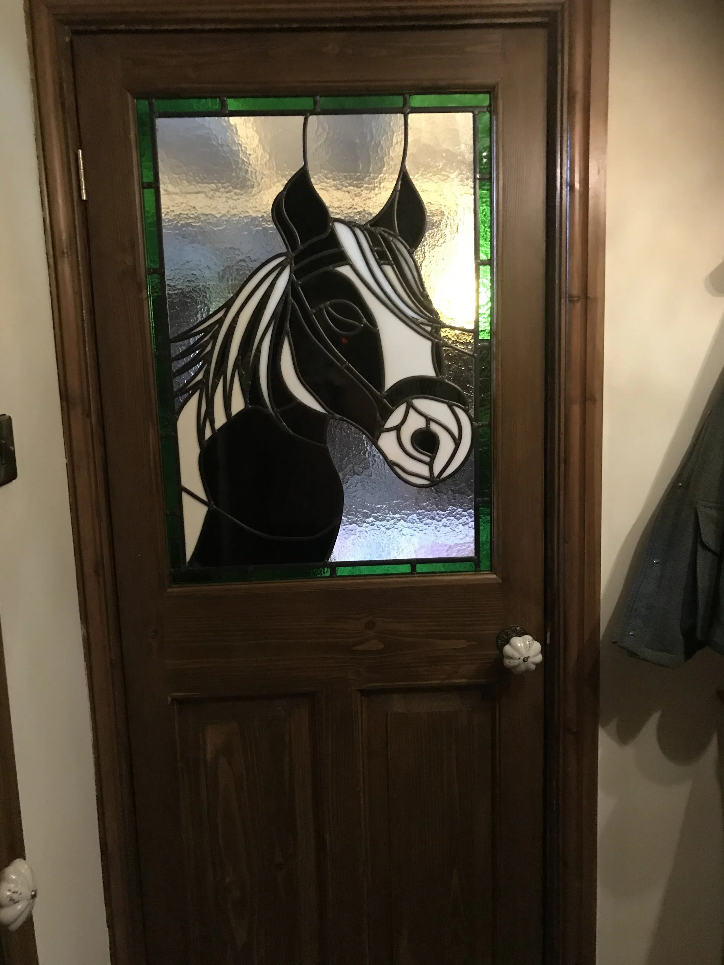 A wooden door with stained glass featuring a horse's head design, framed with green accent glass and textured clear glass.