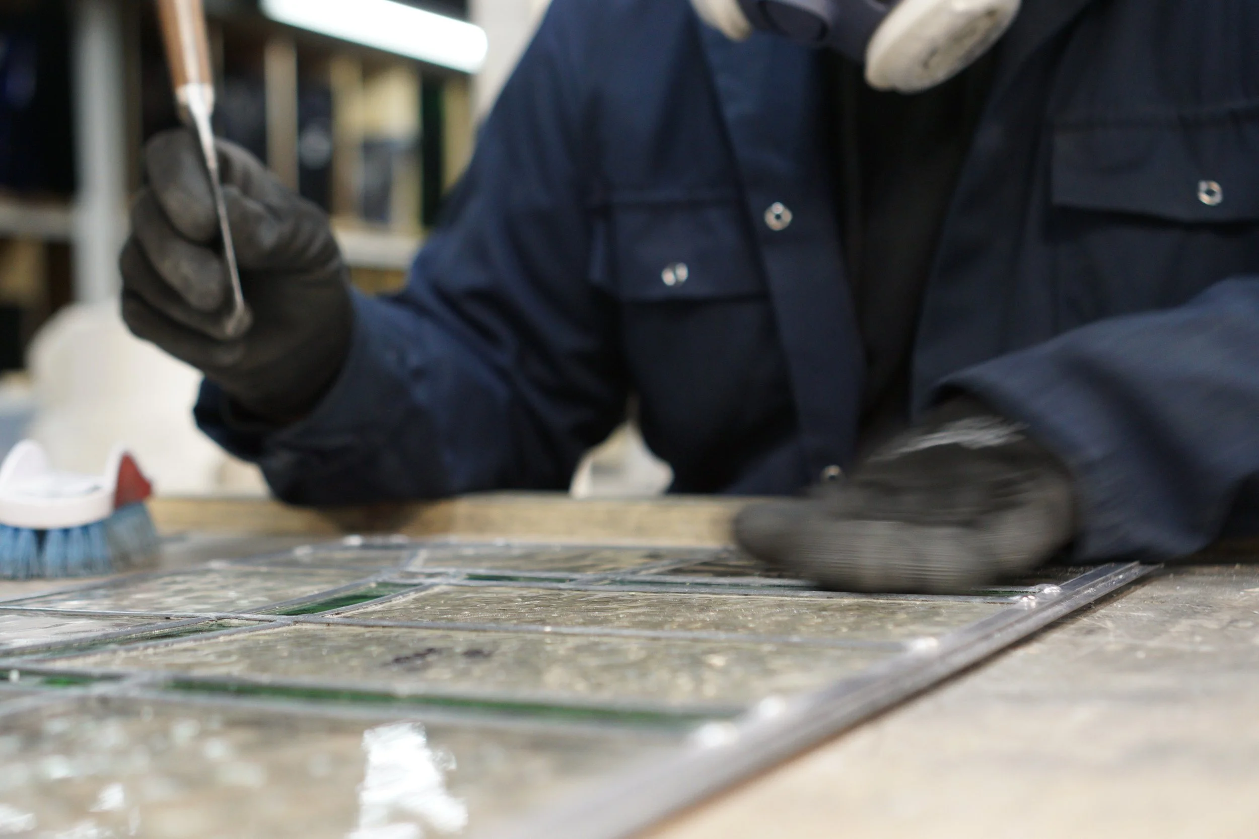 A person wearing a dark blue work jacket and black gloves working with glass tiles, using a tool on the tiles in a workshop.