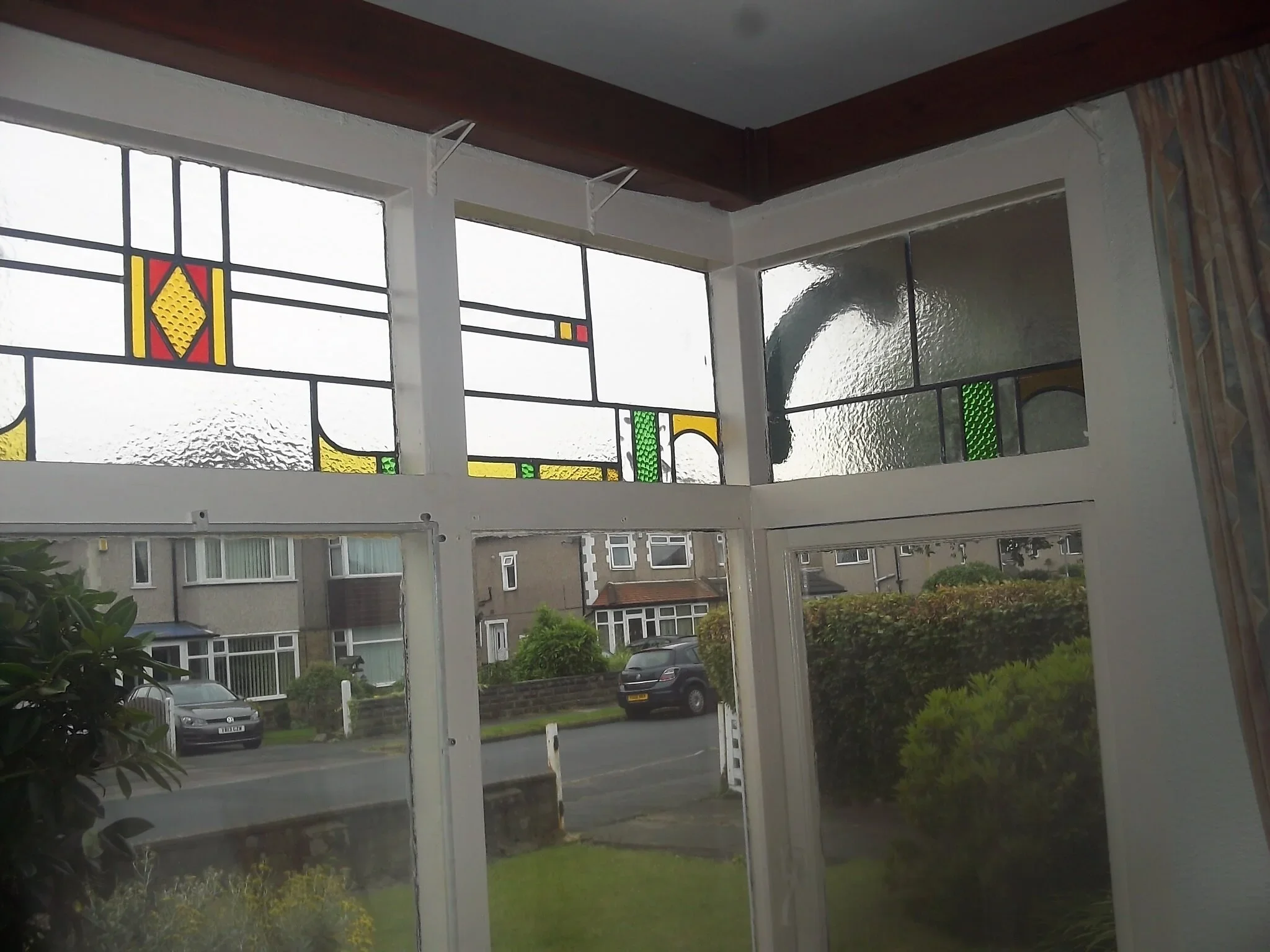 View of a residential neighborhood seen through a window with stained glass design, showing parked cars, houses, and greenery outside on a cloudy day.
