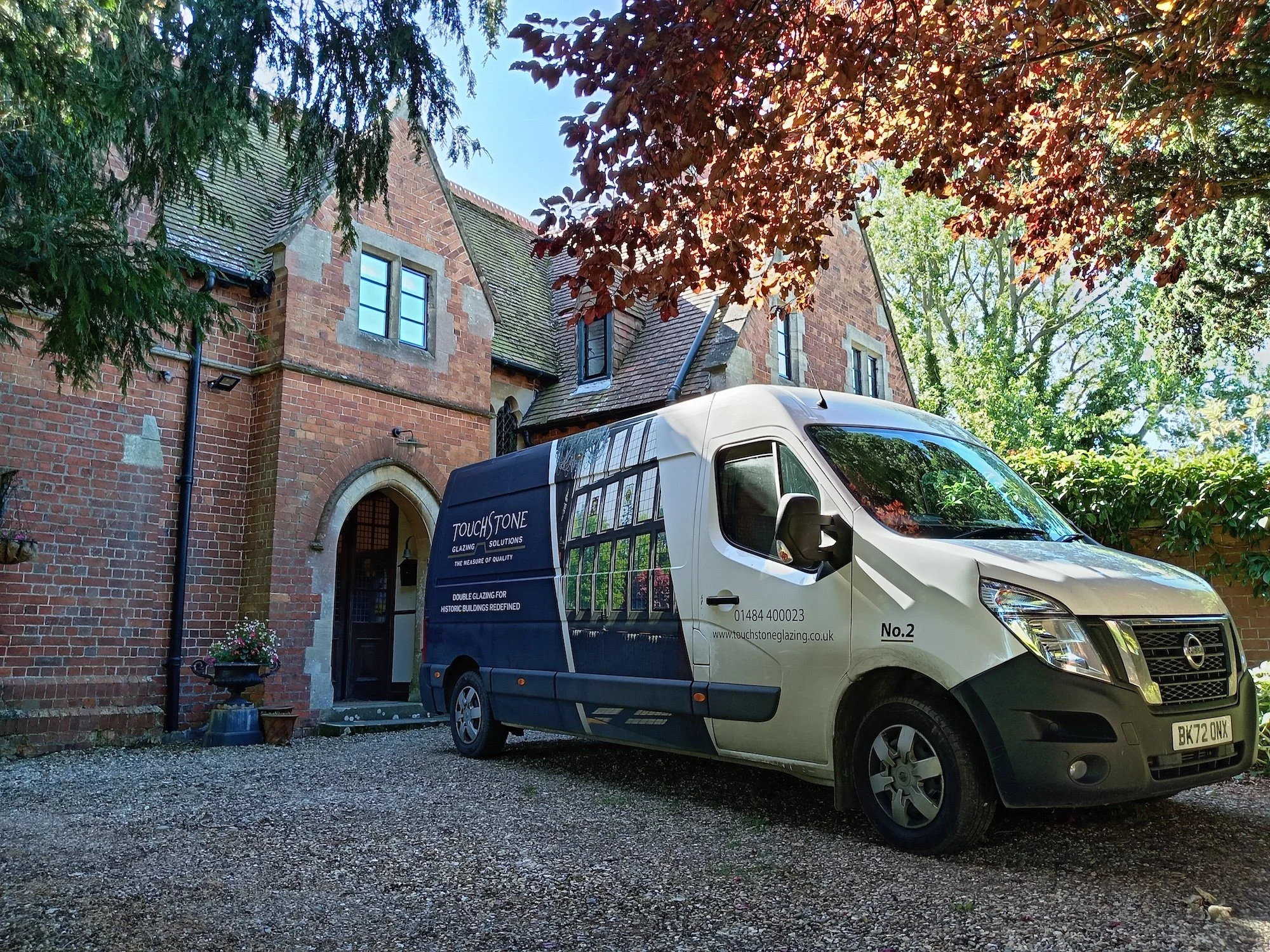A white work van parked in front of a brick house surrounded by trees in a lush garden.