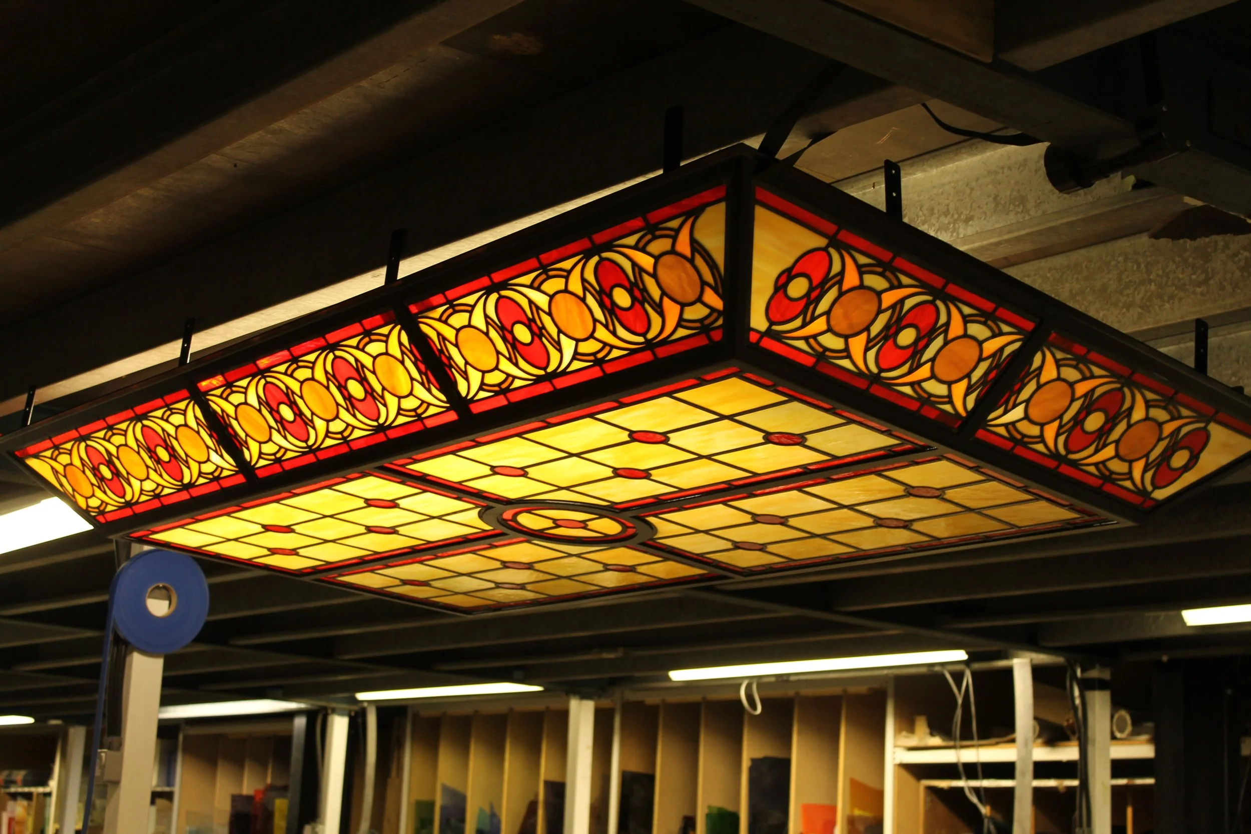 A stained glass ceiling light fixture with a geometric and floral design in yellow, orange, red, and black, installed in an industrial ceiling.