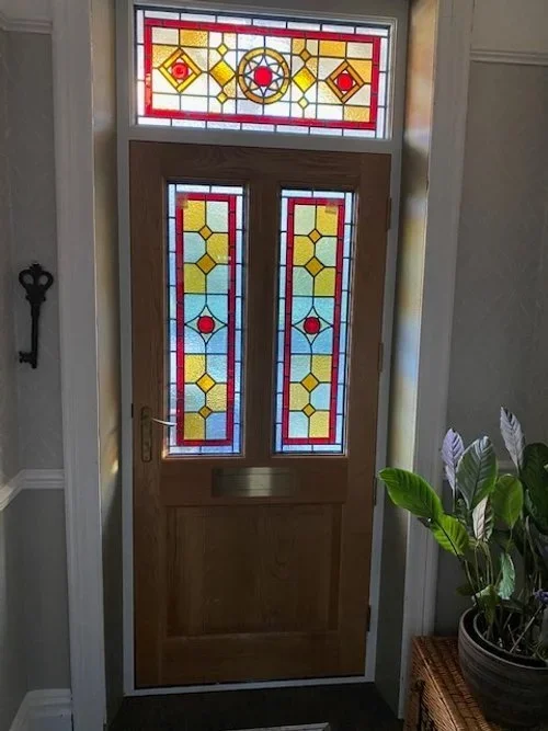 Wooden front door with stained glass panels and a transom window above, featuring a geometric pattern with red, yellow, and blue glass.
