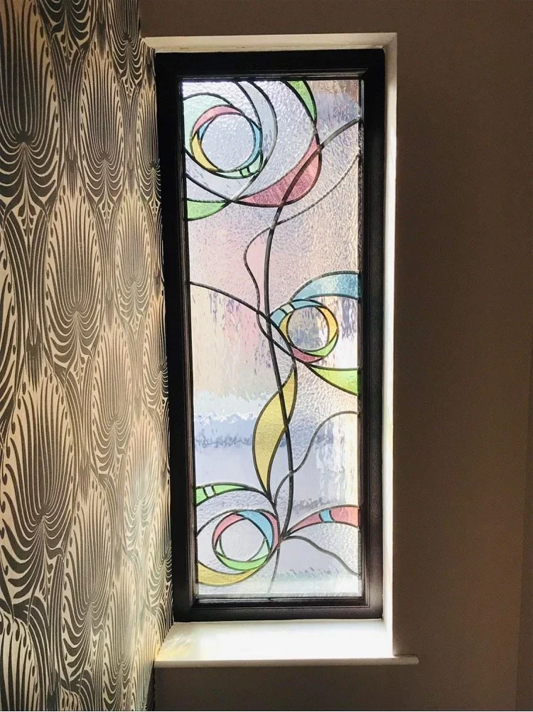 Decorative stained glass window with colorful swirling floral patterns in a black frame, mounted on a wall with a patterned wallpaper.