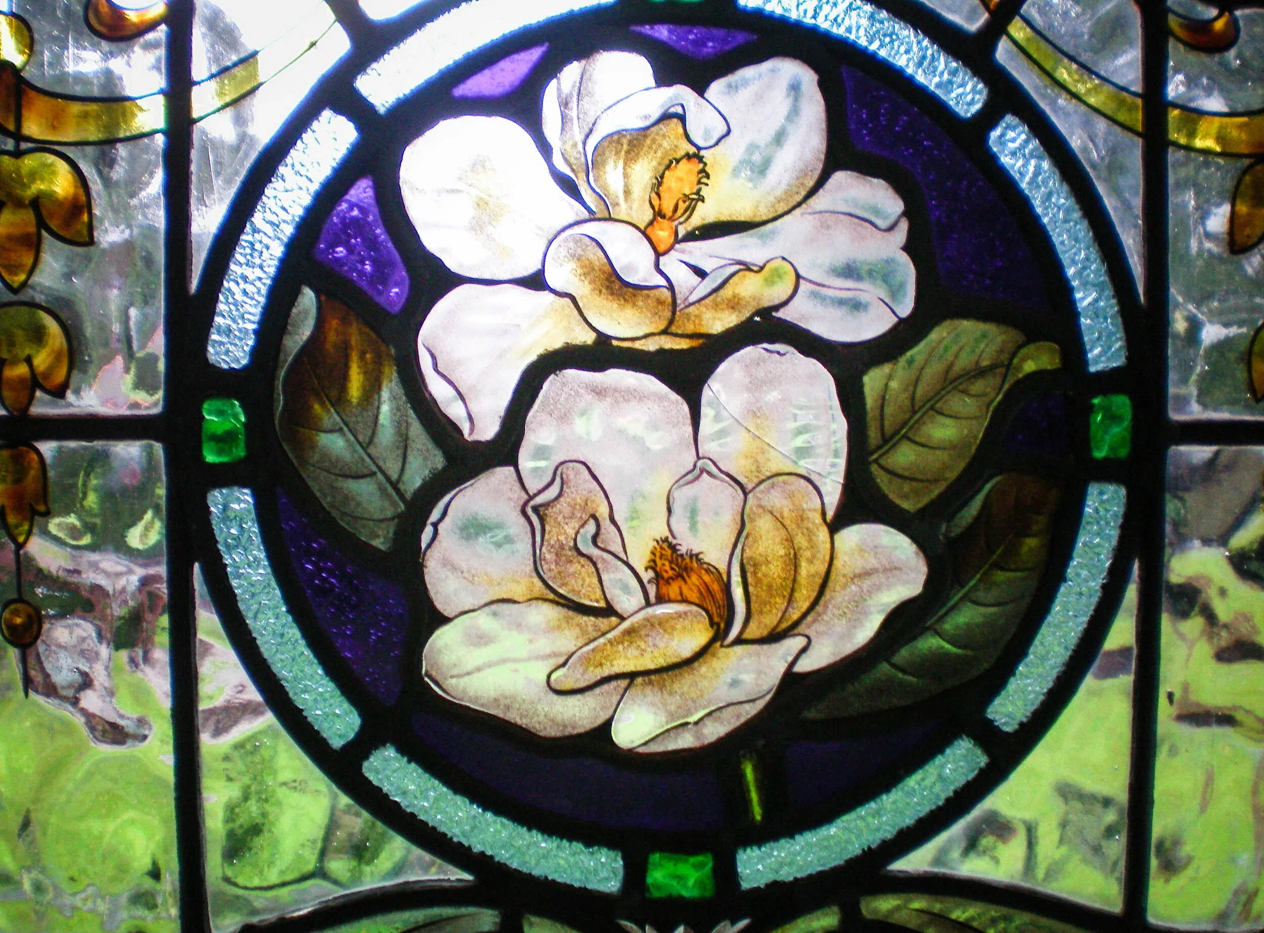 Close-up of a stained glass window featuring a large white flower with orange and purple accents, surrounded by green leaves and decorative glass segments.