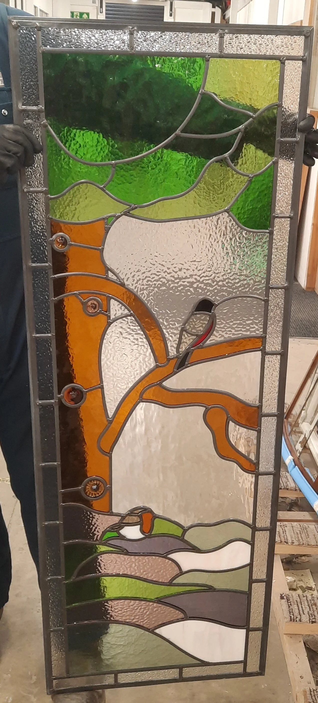 Colorful stained glass window panel featuring abstract design with curved shapes in green, orange, white, brown, black, and purple, set in a metal frame.